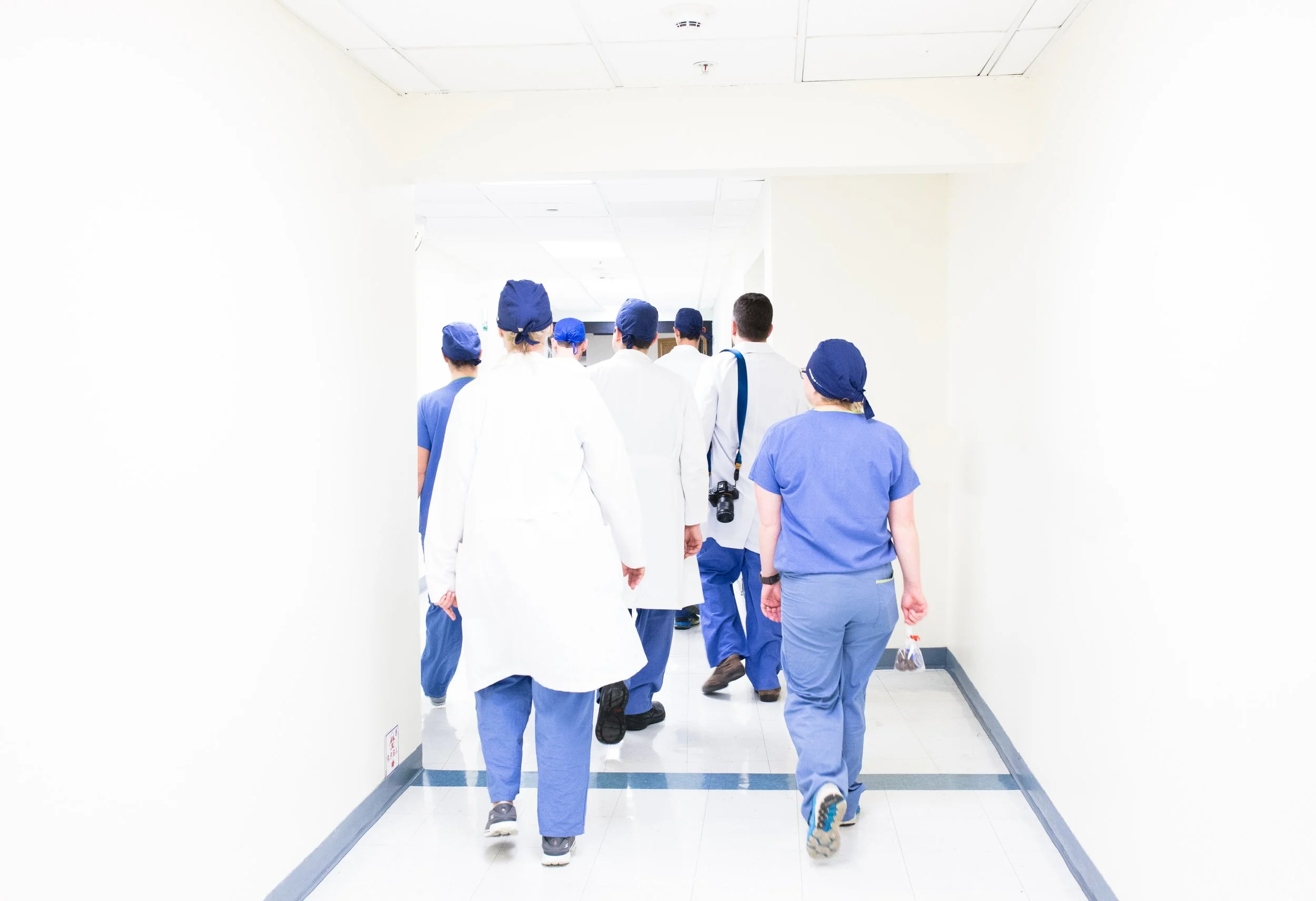 Doctors in white coats and scrub caps with nurses in blue scrubs walking down a white hall into an operating room