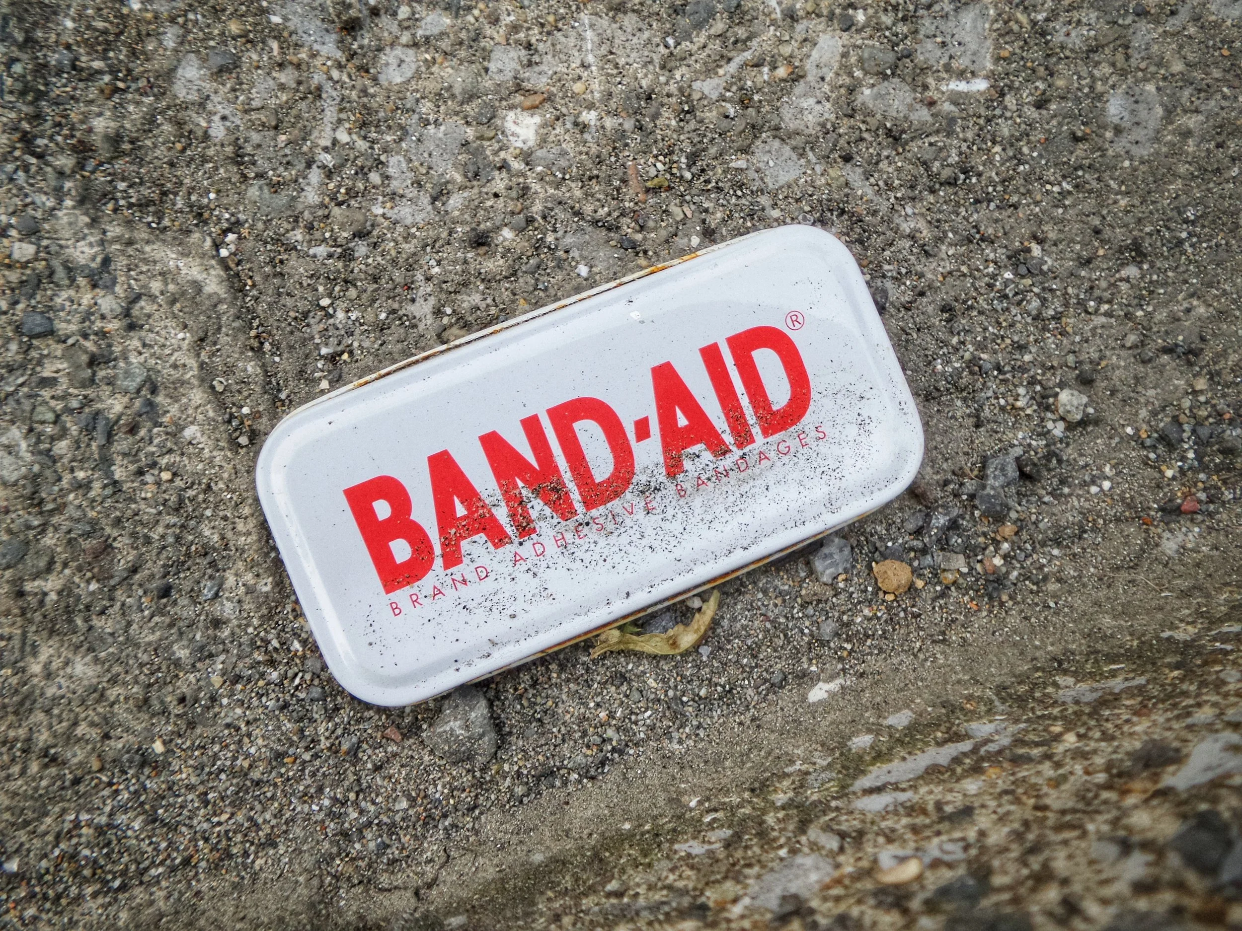 White tin Band-Aid container laying on curb, covered in sand