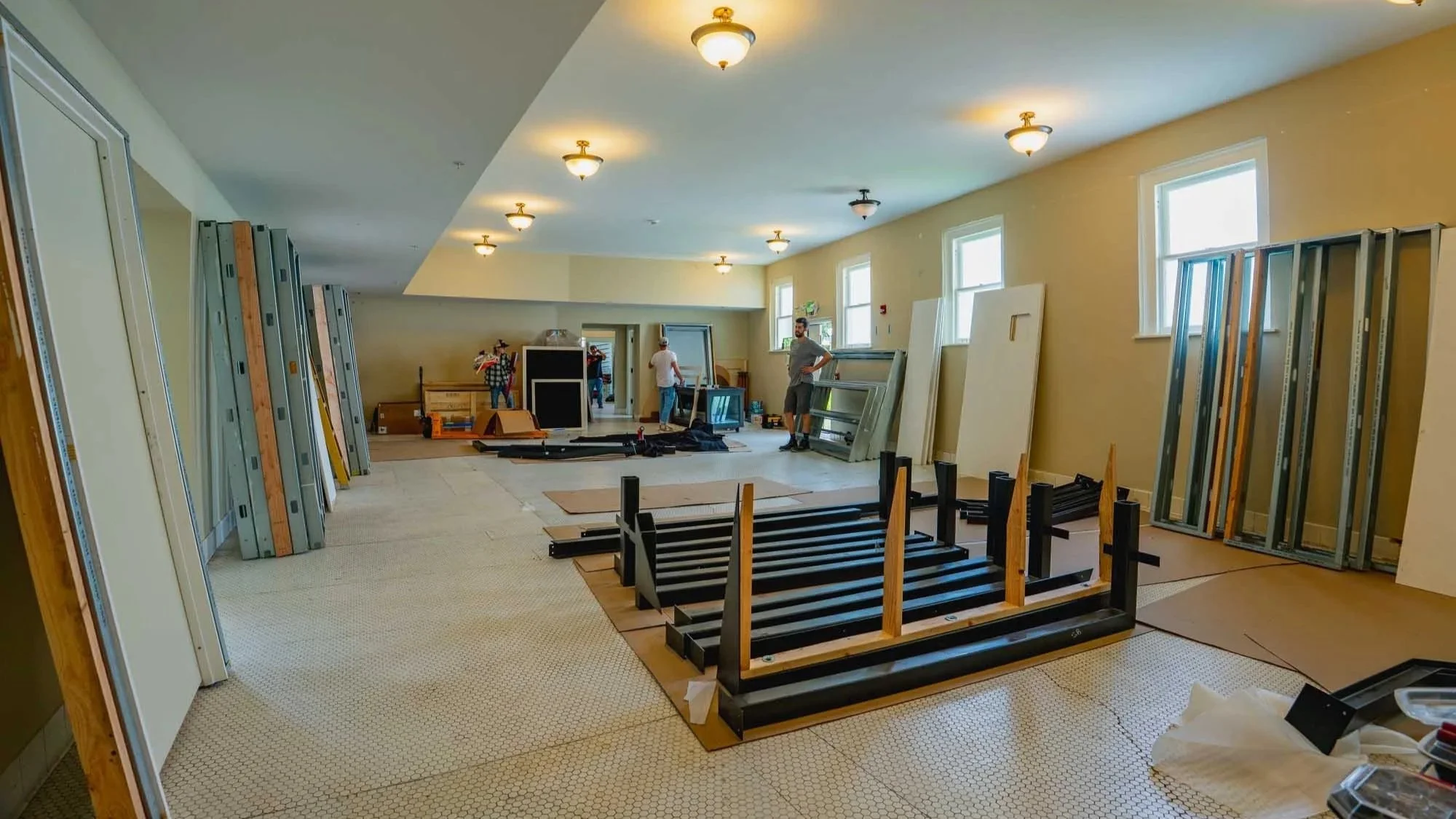 Room being renovated with furniture and supplies against walls, people working in the background, ceiling lights, windows, and construction materials scattered on the floor.