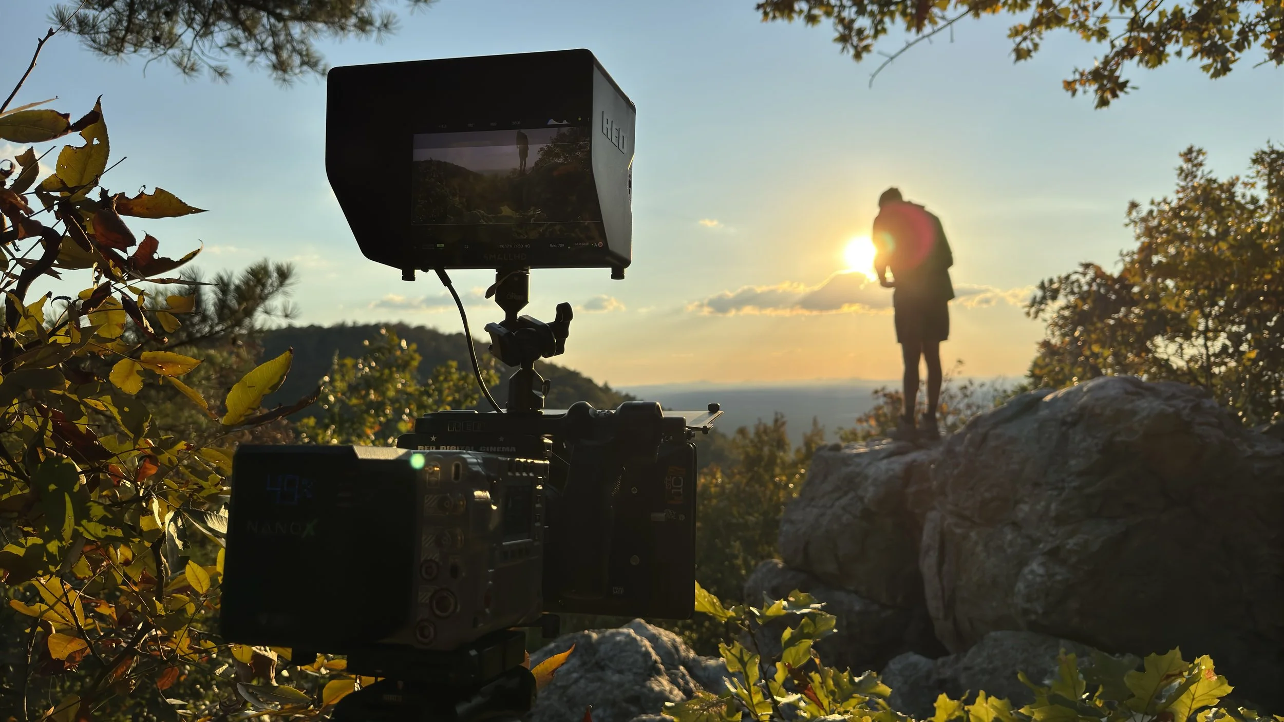 A camera setup capturing a person standing on a rock at sunset, framed by trees and foliage, with the sun in the background.