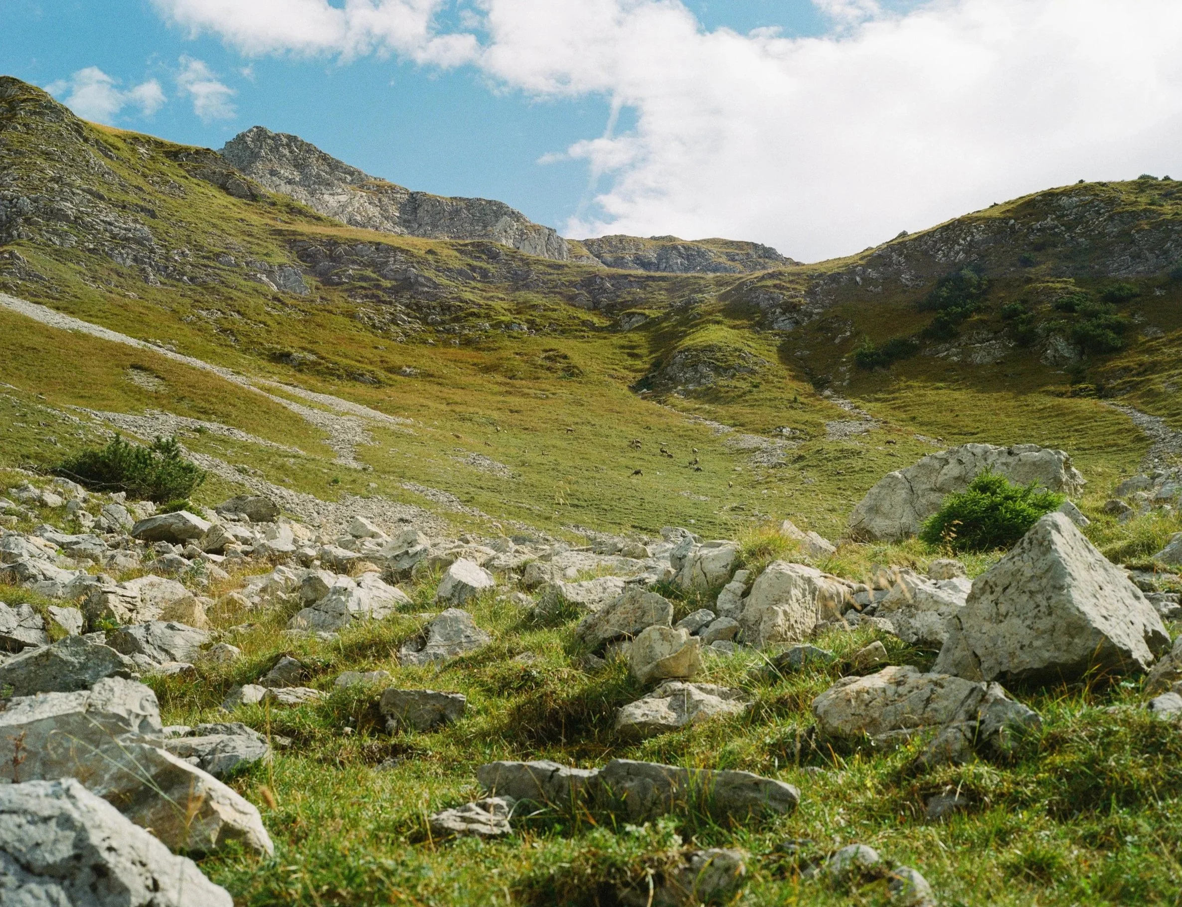 Mountain landscape with rocky slopes, grassy fields, and a partly cloudy sky.