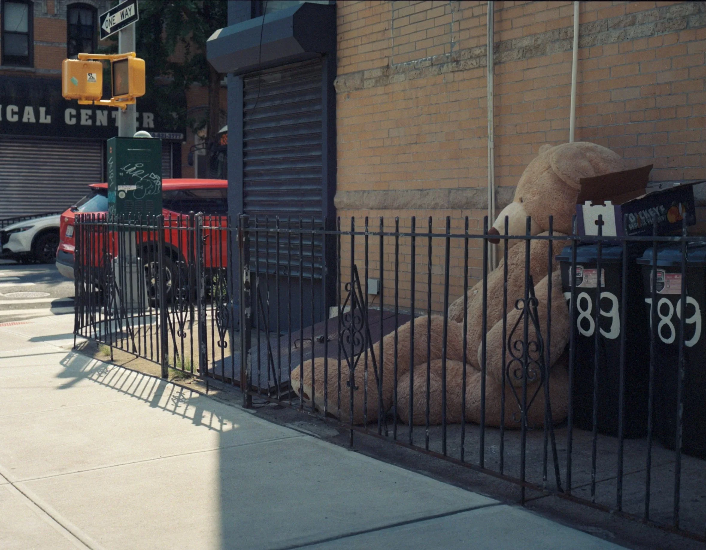 A city sidewalk with a brick building, a black metal fence, a large stuffed bear leaning against the building, a cardboard box with food branding, a stopped red car, and a traffic light, street sign, and utility pole.