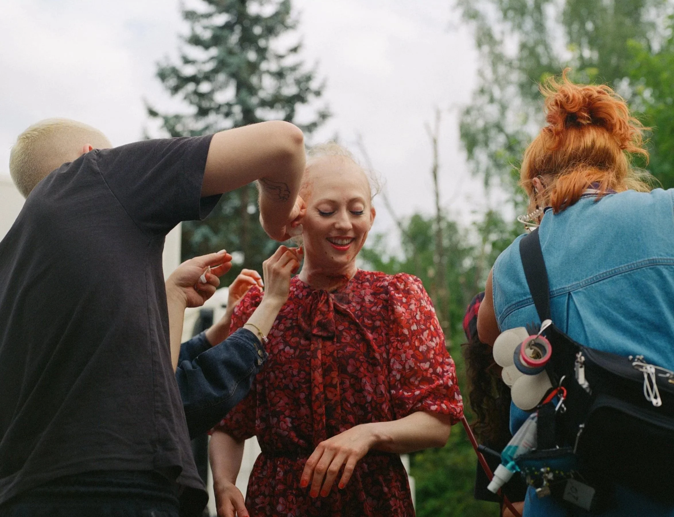 Group of people outdoors, including a woman, wearing a patterned red shirt, receiving makeup services from others.