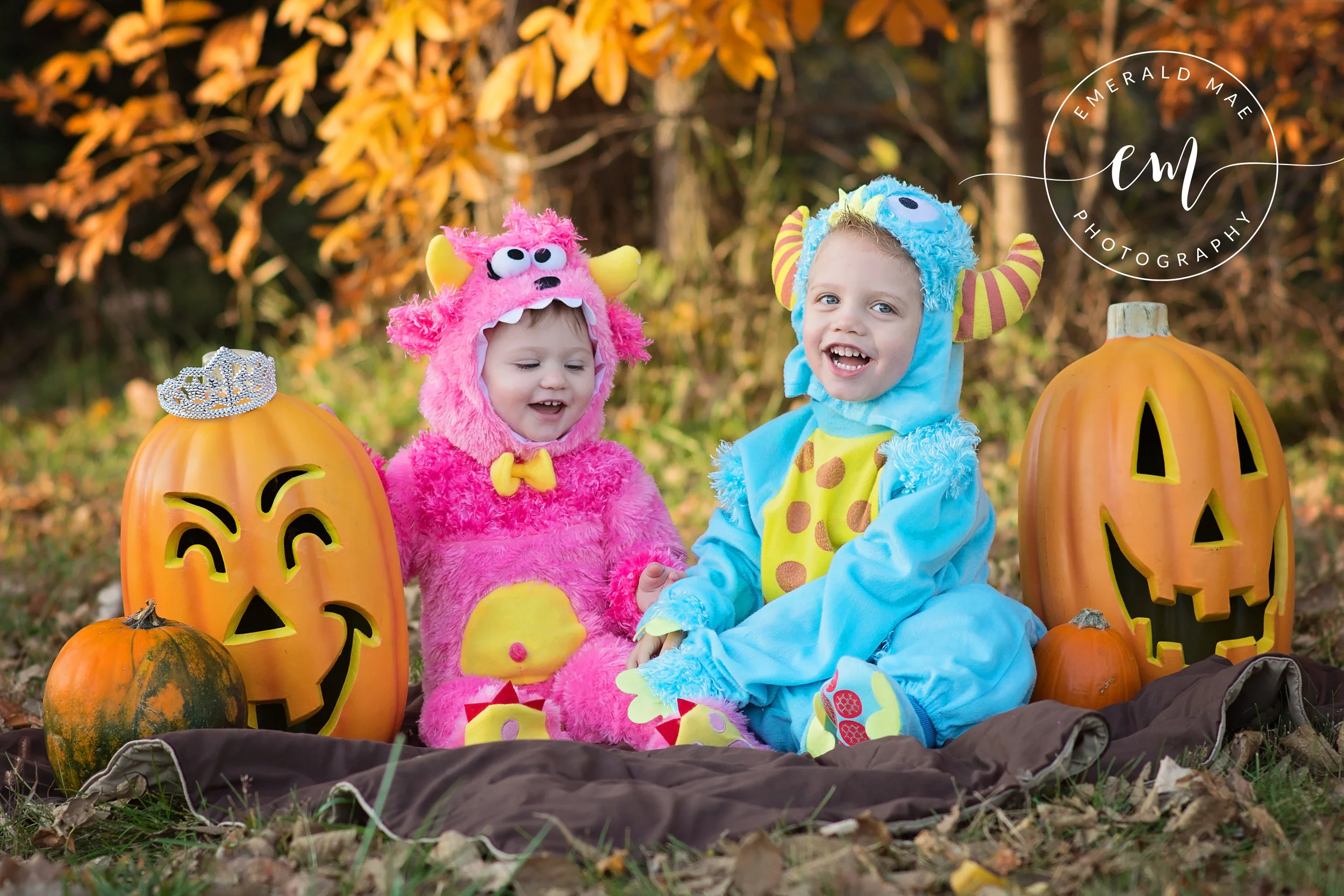 Two children in colorful monster costumes sitting outdoors with carved Halloween pumpkins, autumn leaves, and a blanket.
