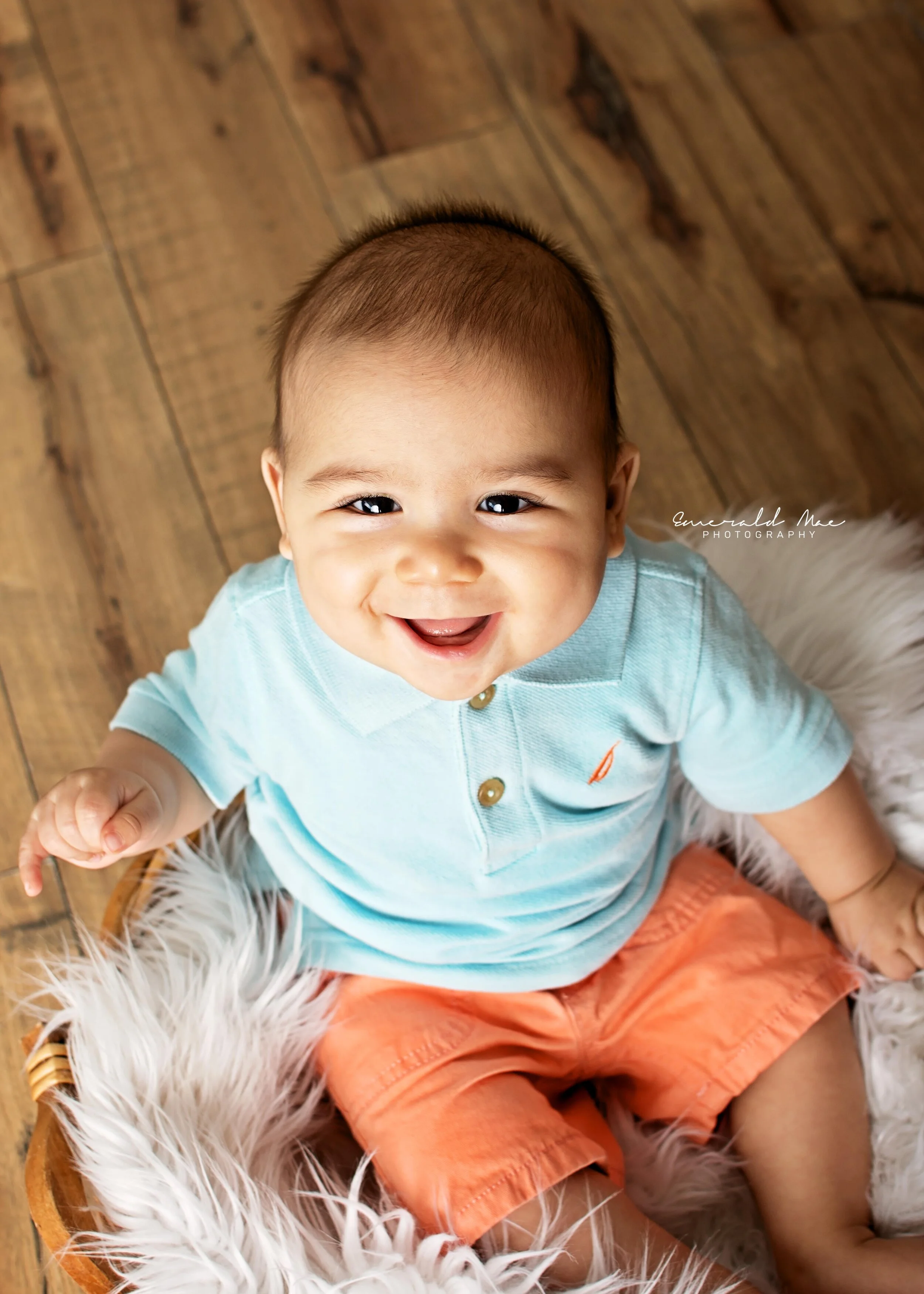  A smiling baby with short hair sits on a plush white blanket in a wicker basket. The baby is wearing a light blue polo shirt with an orange sailboat emblem and matching orange shorts. The background features wooden flooring. The portrait was capture