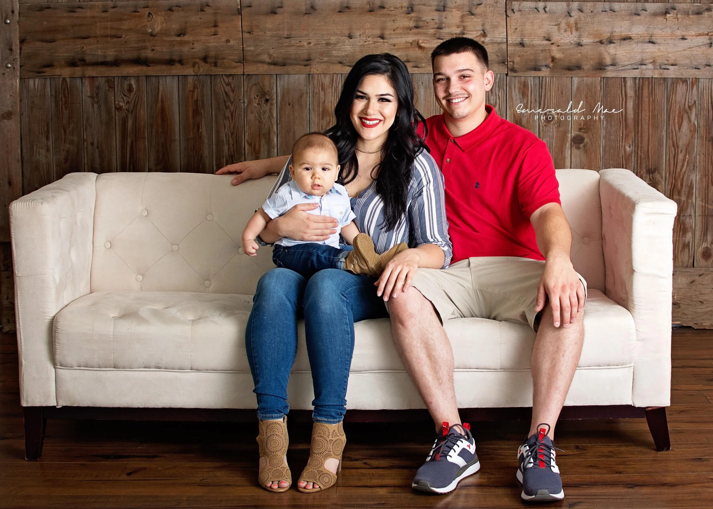  A family of three sits on a beige couch in front of a wooden wall. The mother, holding a baby, is wearing a striped top and jeans; the father is wearing a red polo shirt and khaki shorts. The baby is dressed in a light shirt and blue pants. They all