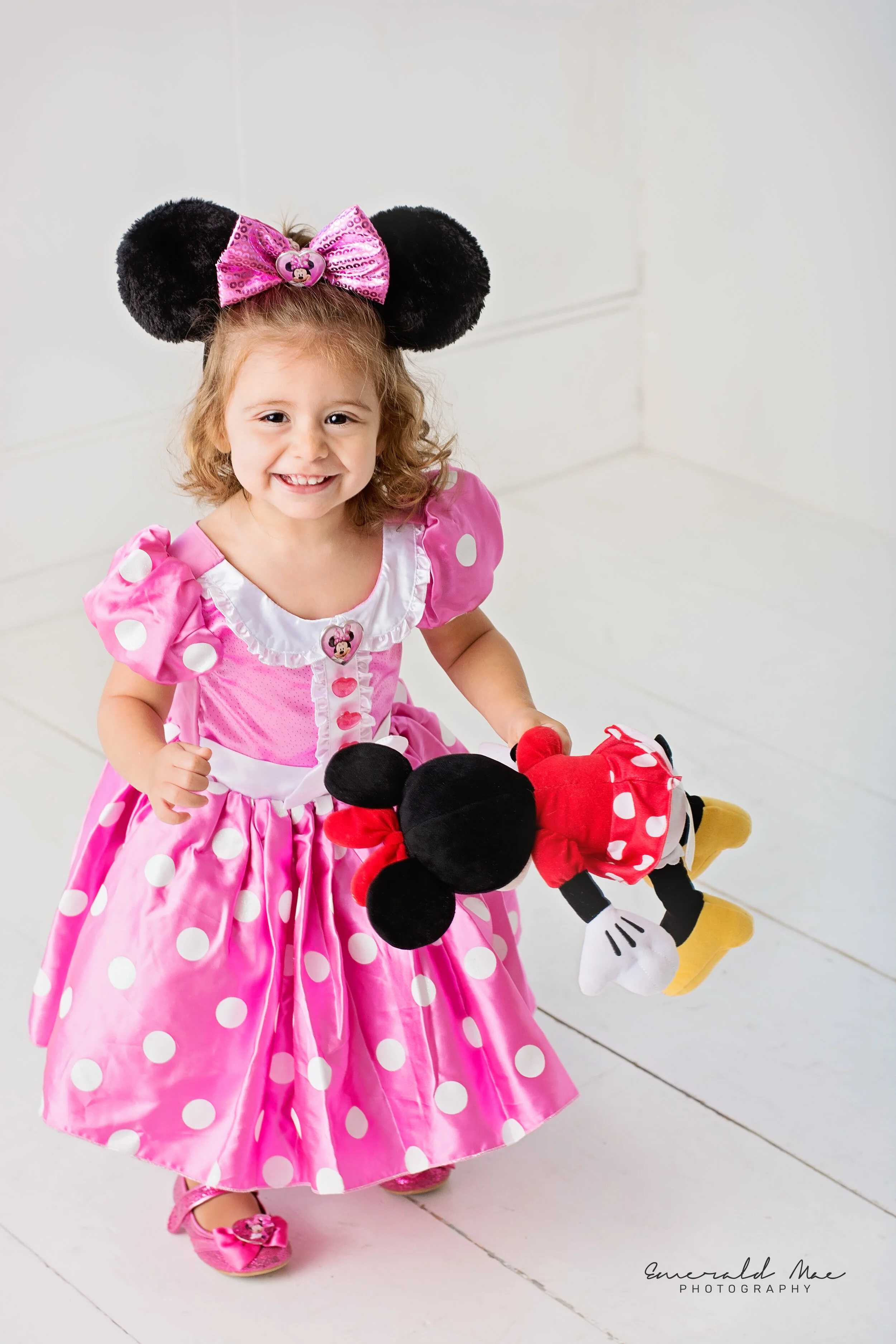  A young girl wearing a pink polka dot dress and Minnie Mouse ears smiles while holding a Minnie Mouse plush toy during her birthday photo session by Emerald Mae Photography. She stands on a white floor with a plain white background at the photograph