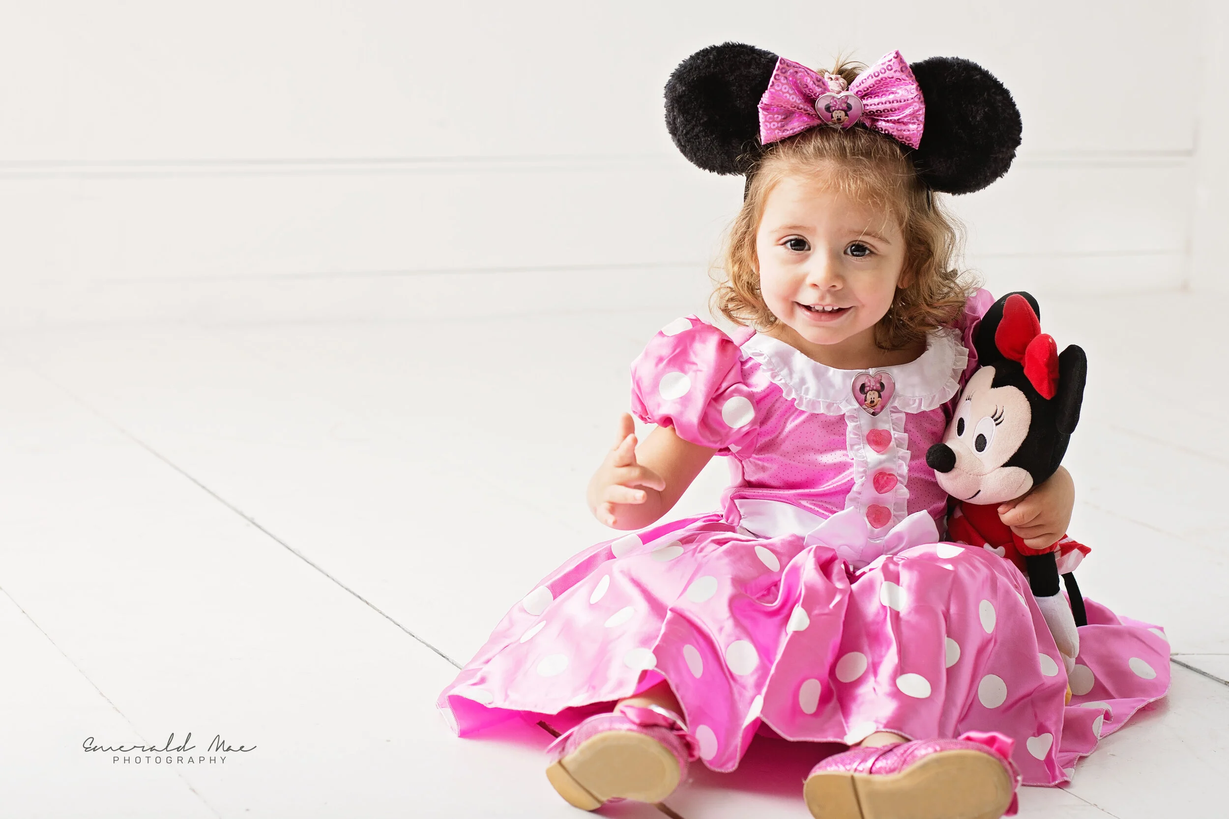  Omaha area family photographer, Emerald Mae Photography, captures a young girl in a pink polka-dotted dress, large Minnie Mouse ears sitting on a white floor, smiling at the camera and holding a Minnie Mouse stuffed toy for her birthday photo sessio