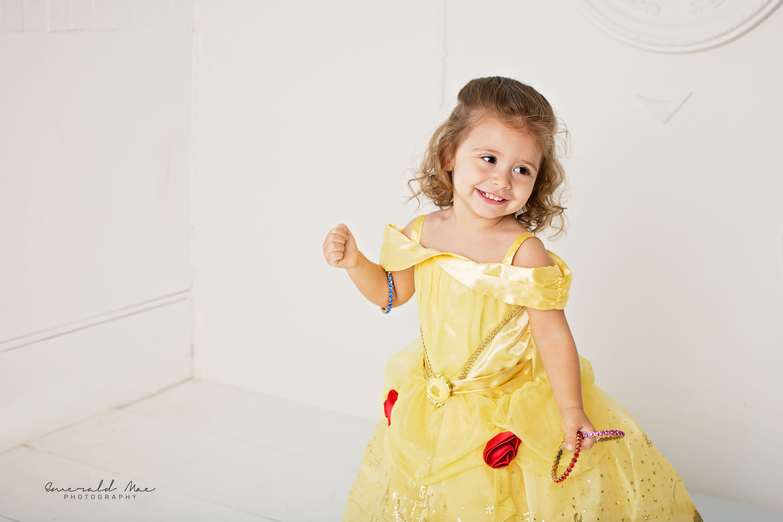  A young child is smiling and posing for her birthday in a bright yellow, Princess Belle dress with red roses and glitter details. The child has curly hair and is wearing colorful bracelets. Captured by Omaha photographer Breanna DeWitt of Emerald Ma