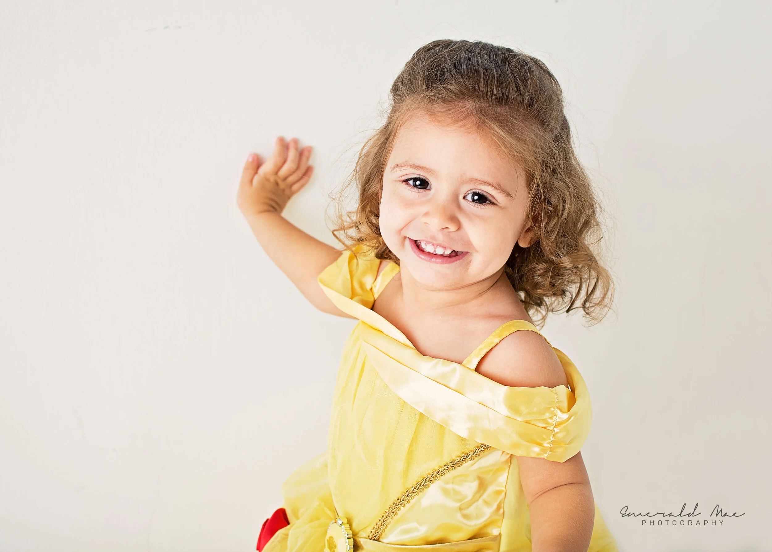  A smiling young child with wavy brown hair dressed as her favorite Disney Princess, Belle from Beauty and the Beast, poses against a plain white background for her birthday photo session. The child has one arm raised and their hand touching the wall