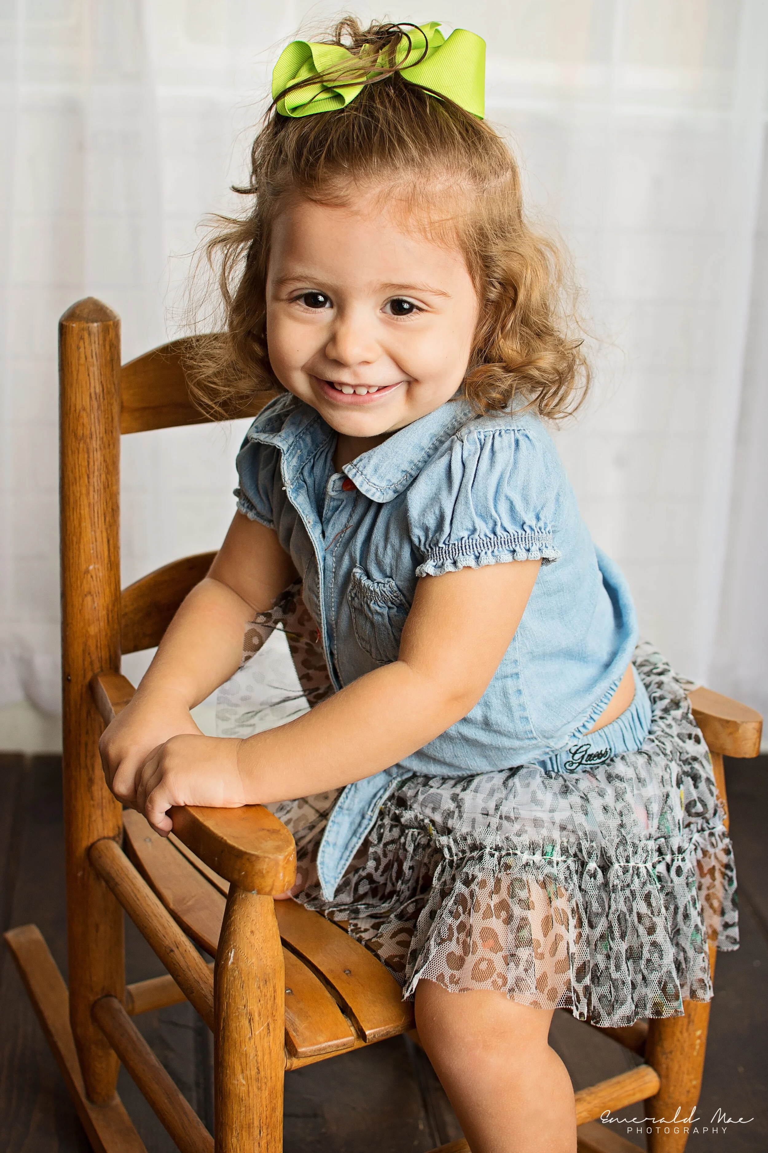  A young girl with curly hair, wearing a denim shirt and a leopard print skirt, sits smiling on a wooden rocking chair. She has a lime green bow in her hair and is looking at the camera with a cheerful expression. Perfect for family photography, this
