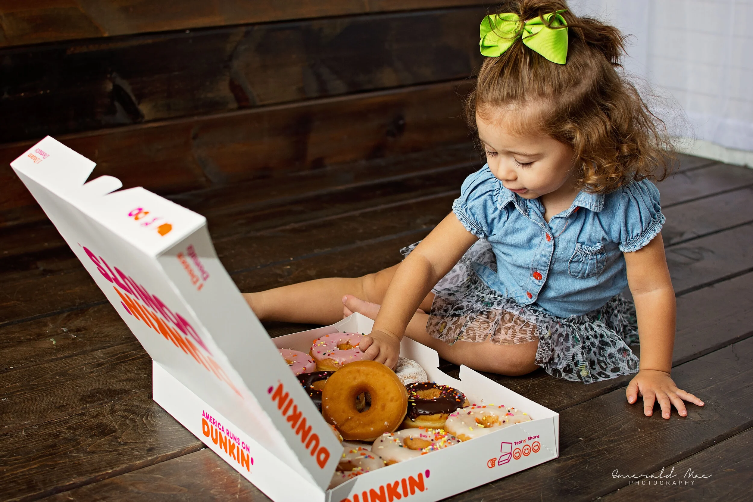  A young girl with a green bow in her hair, wearing a denim shirt and leopard print skirt, sits on the floor and reaches into a Dunkin' Donuts box containing assorted donuts. The background features dark wooden flooring—perfect for children's birthda