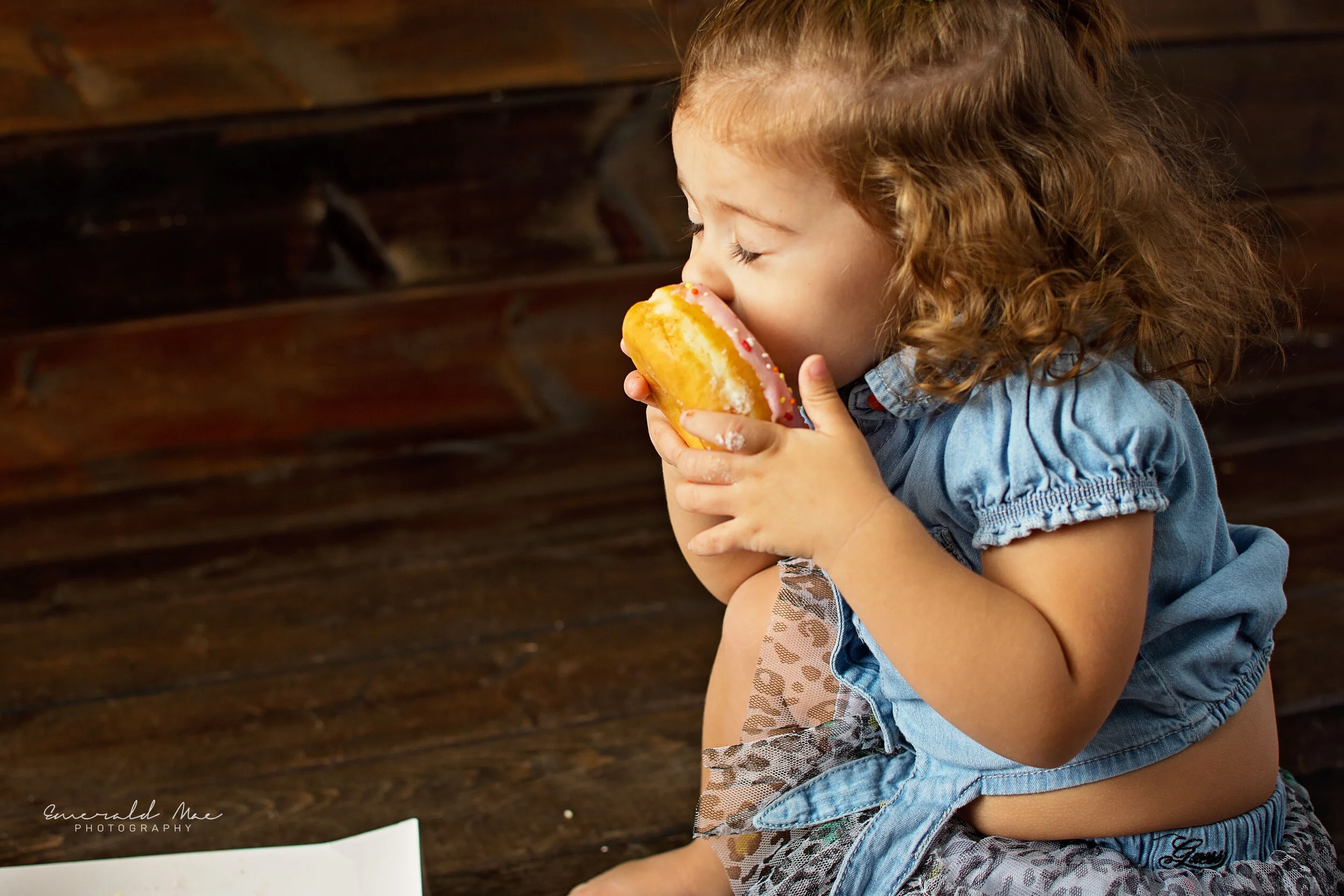  A young child with shoulder-length curly hair is sitting on the floor and taking a big bite out of a donut. The child is wearing a light blue shirt and patterned leggings, perfectly capturing the charm of children's photography against the rustic wo