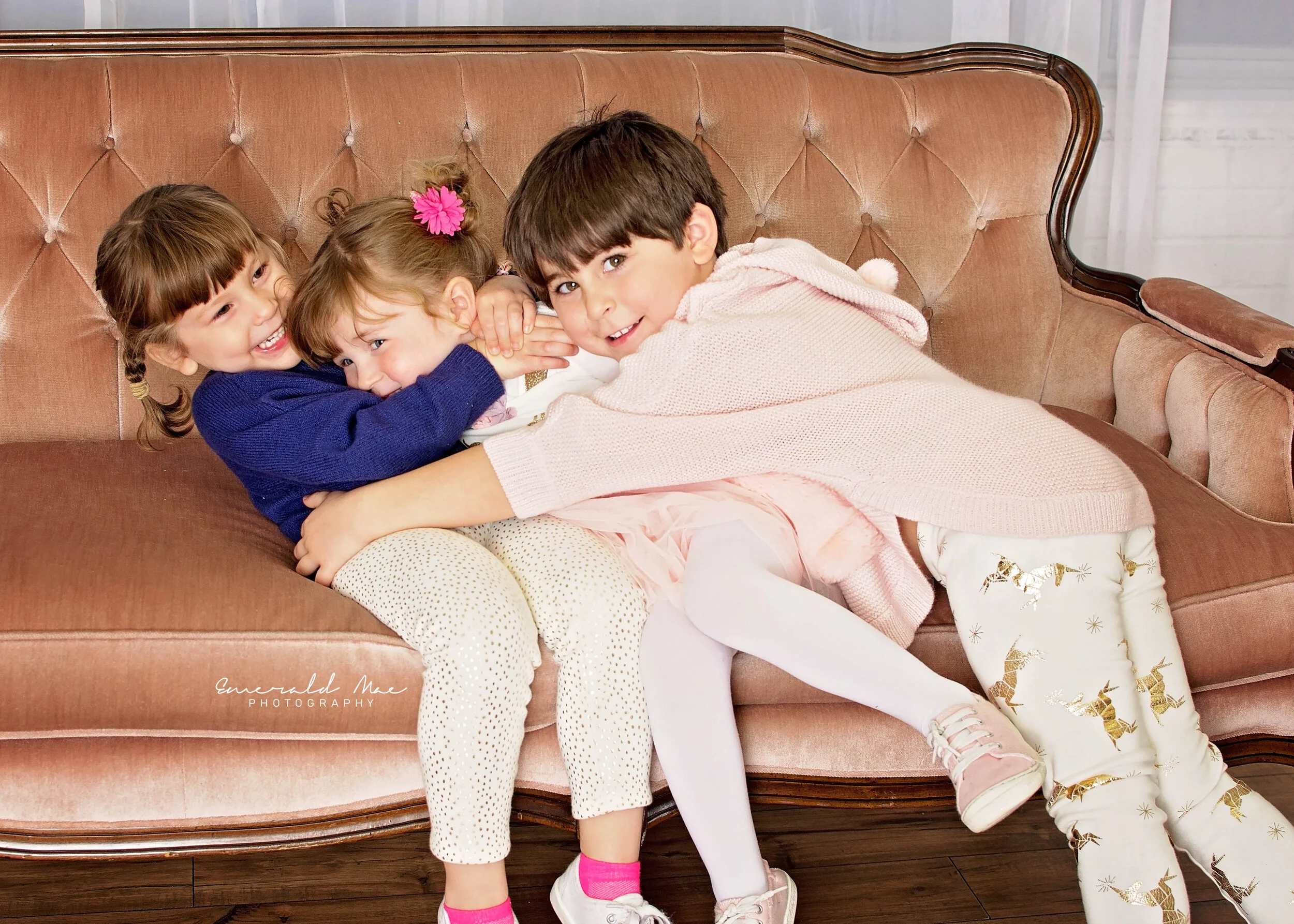  Three children are sitting on a plush, vintage-style couch. Two girls, with the one in blue hugging the other in white, share a laugh. The boy in a light pink sweater and patterned pants joins the embrace, smiling warmly. This heartwarming moment of