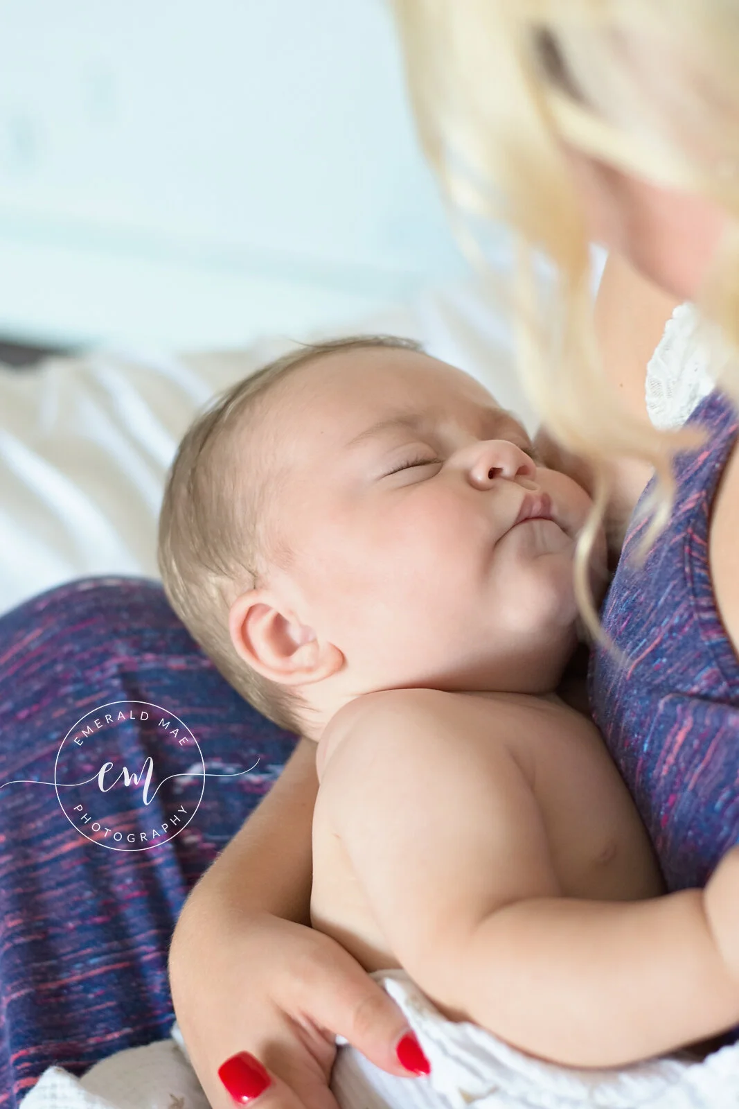  A close-up image shows a baby sleeping peacefully while being cradled in the arms of their mother, whose face and hair is partially visible. The mother's bright red nails and purple patterned shirt stand out. The watermark reads "Emerald Mae Photogr