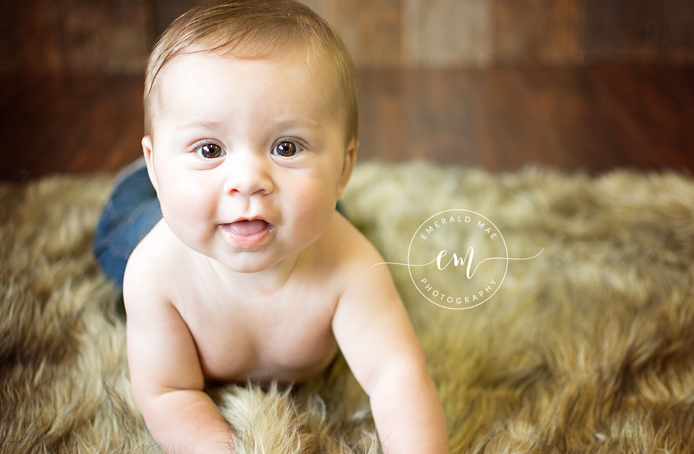  A baby with short light brown hair, wearing no shirt, is lying on a furry rug and looking at the camera. The background of this charming studio photography features a wooden floor and wall, with a watermark reading "Emerald Mae Photography", an Omah