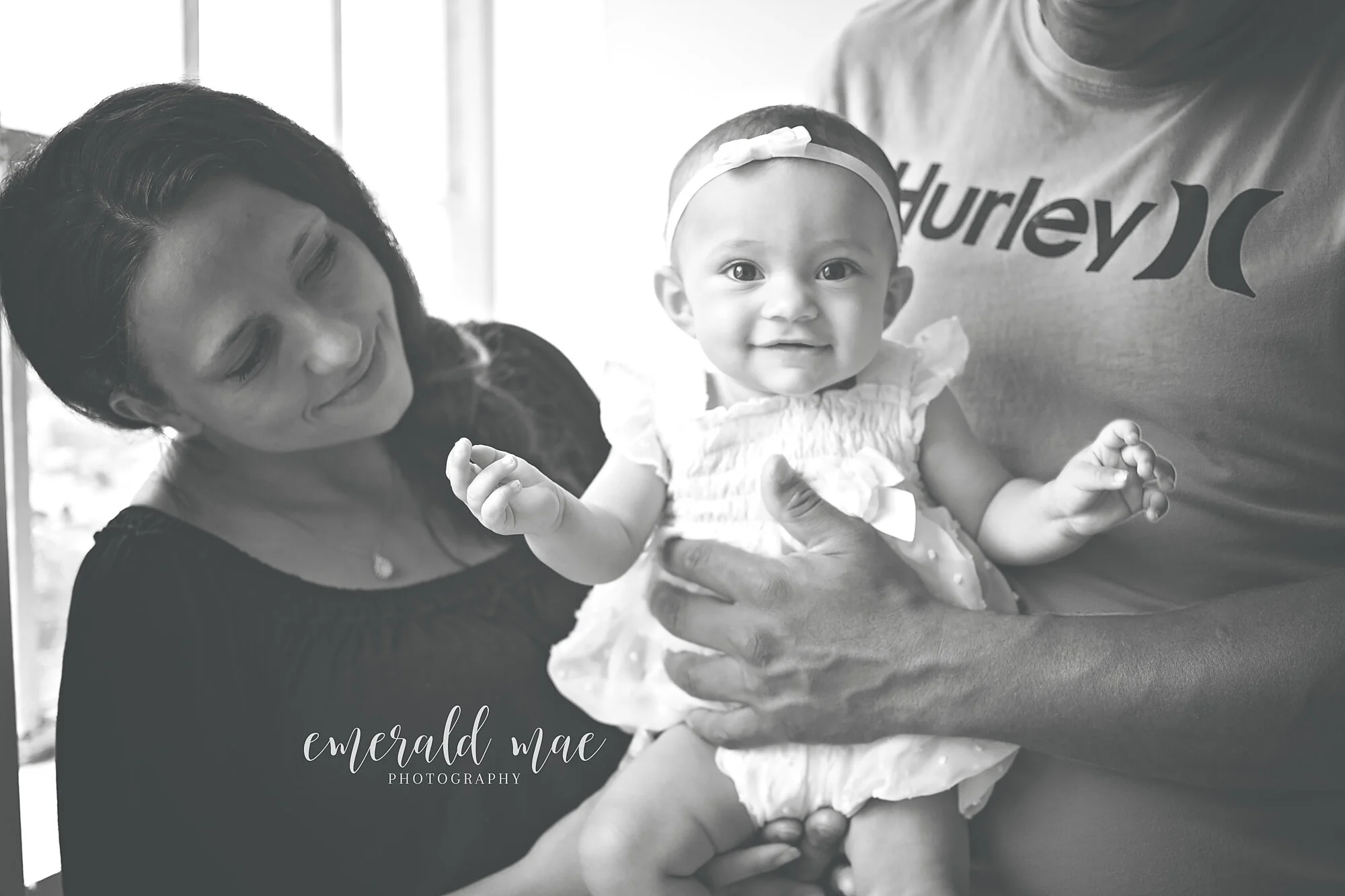  A black and white photo shows a smiling baby in a white dress and headband held by Dad wearing a Hurley T-shirt. Mom looks on lovingly from the left as the baby looks straight at the camera. This heartwarming and candid moment was captured by Emeral