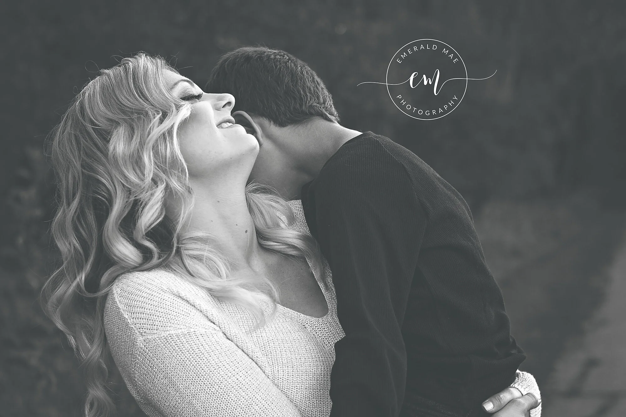  A grayscale image from a family photo session in Omaha, of a smiling woman with wavy hair wearing a knitted sweater. She is holding her son who is nuzzling into her neck. 