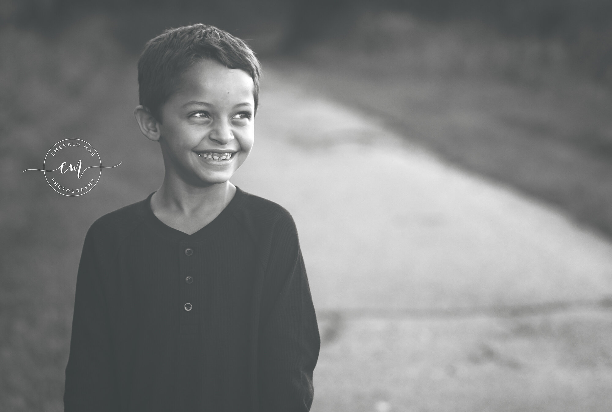  A black and white photo of a young boy standing outdoors on a path. He is smiling and looking to the right, wearing a long-sleeved buttoned shirt. The background is blurred, focusing attention on the child’s joyful expression. 