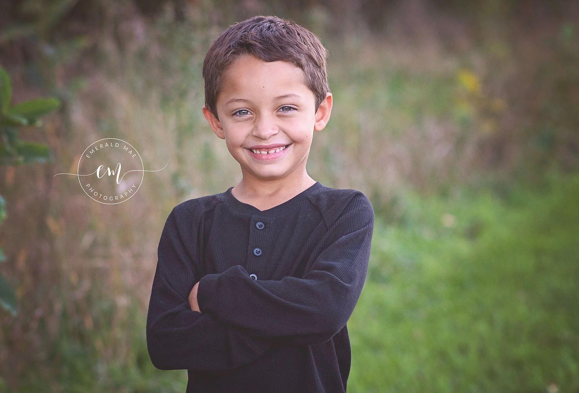  A young boy with short brown hair poses with his arms crossed and smiles at the camera for a family photo session. He is wearing a black long-sleeved shirt and is outdoors with green grass and blurred foliage in the background. The Emerald Mae Photo