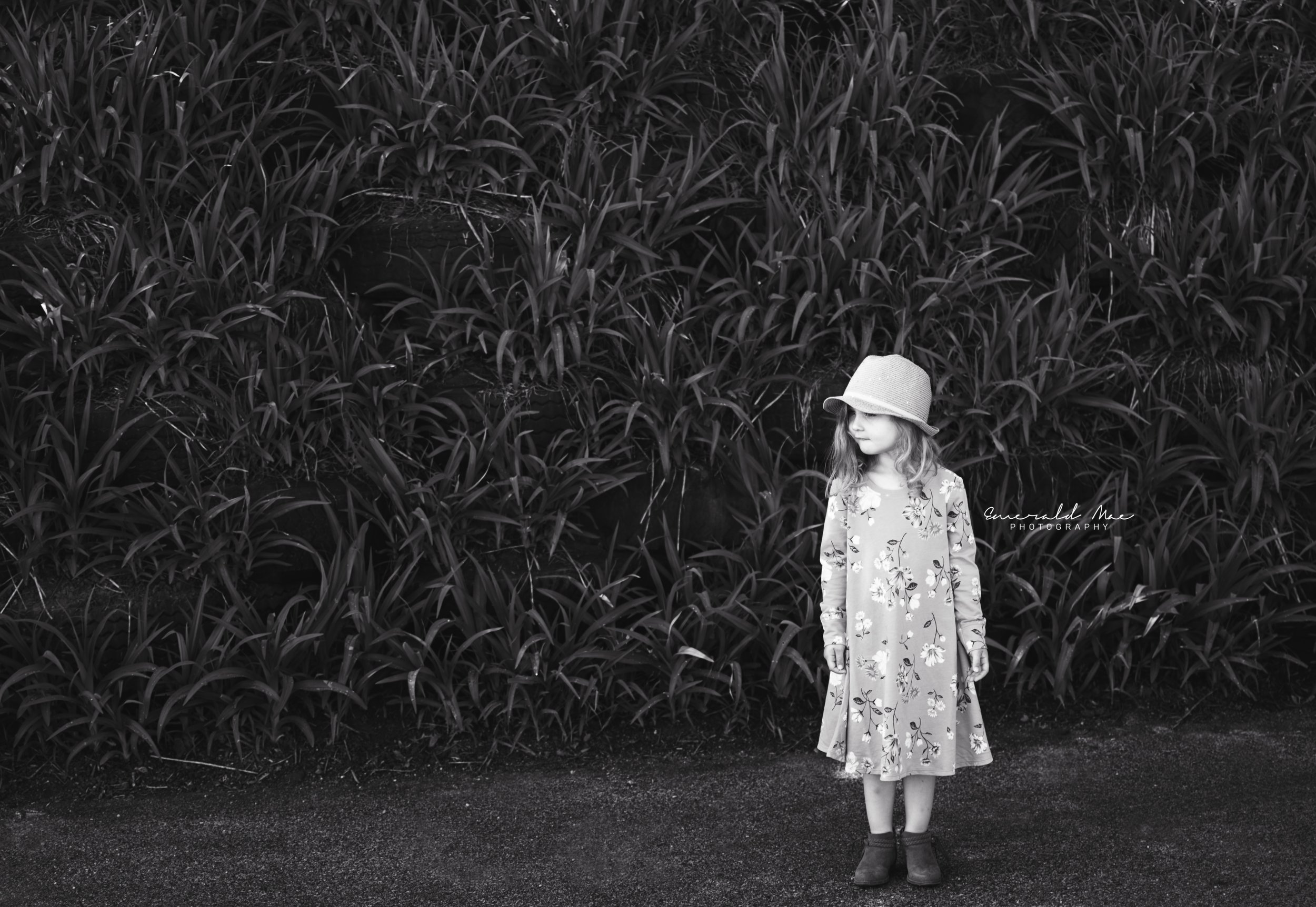  A young girl in a floral dress and hat stands in front of dense foliage, looking to her right. Captured by Emerald Mae Photography in Omaha Nebraska, the image conveys an innocent charm in timeless black and white. 