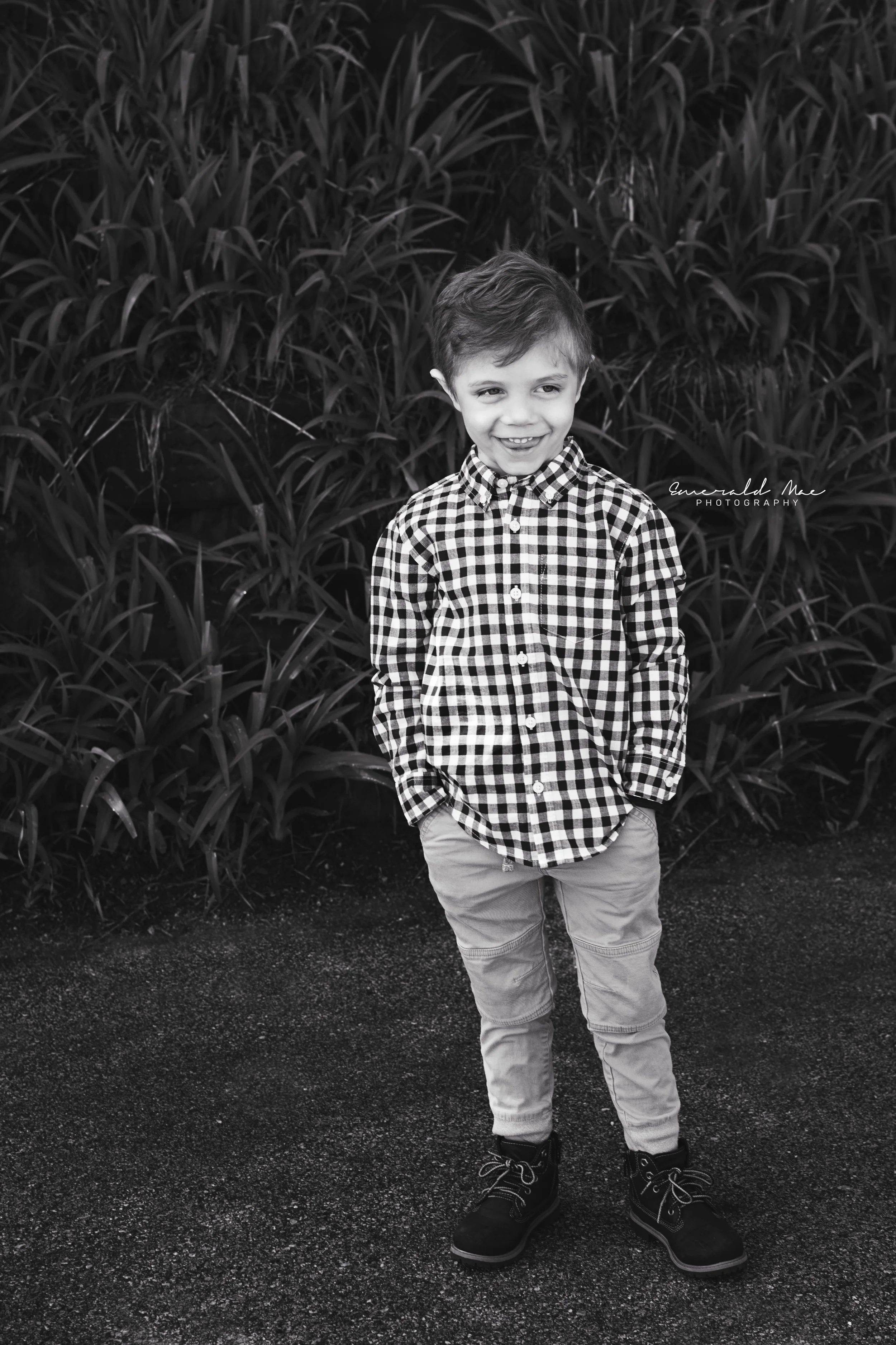  A young child with short hair is standing outdoors on a paved path with hands in pockets. The child is wearing a checkered shirt, light-colored pants, and dark shoes, smiling at the camera. Tall grass or plants form the background in this black-and-