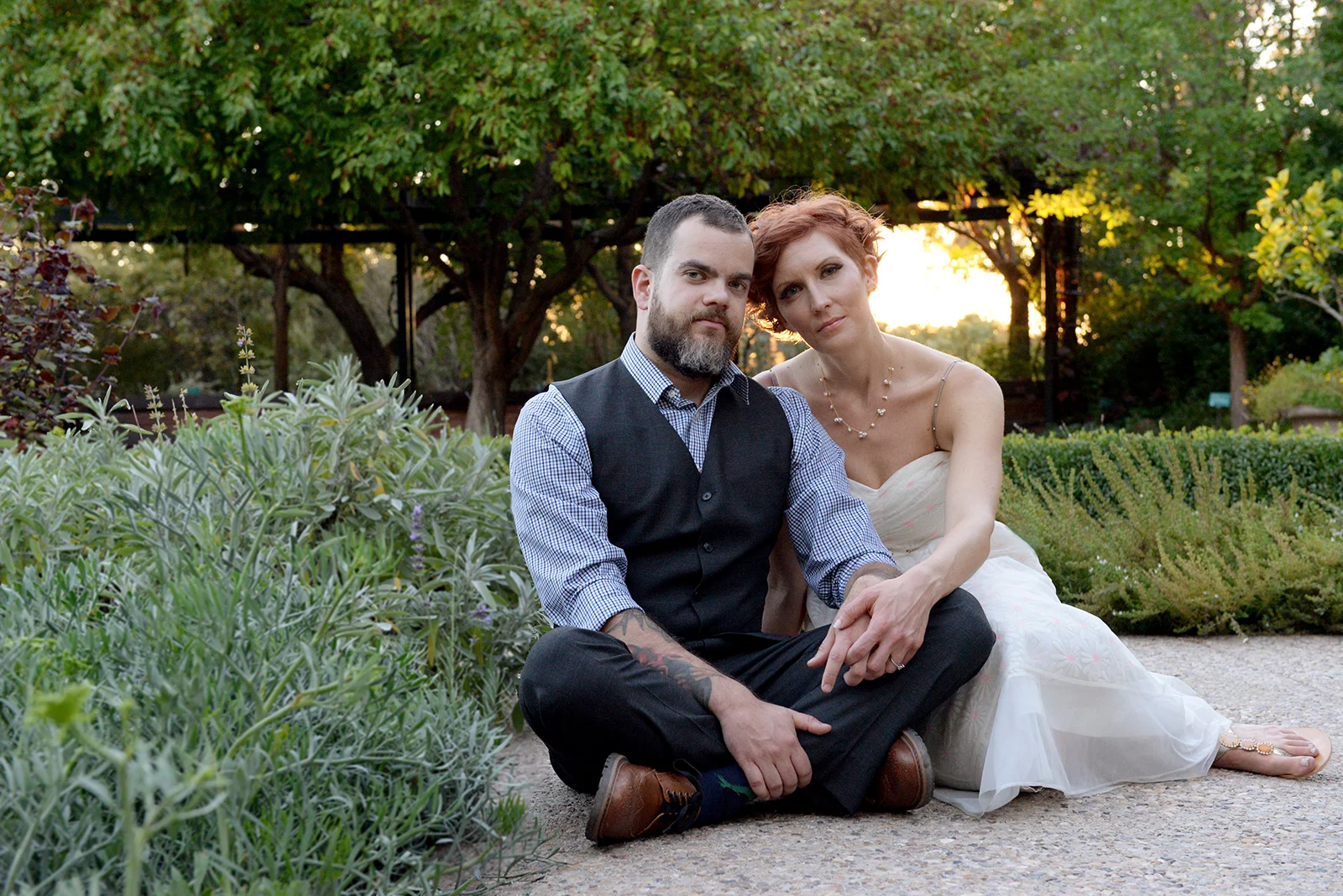 A bride and groom sit together on the ground in a lush desert garden with hedges and trees in the evening light.