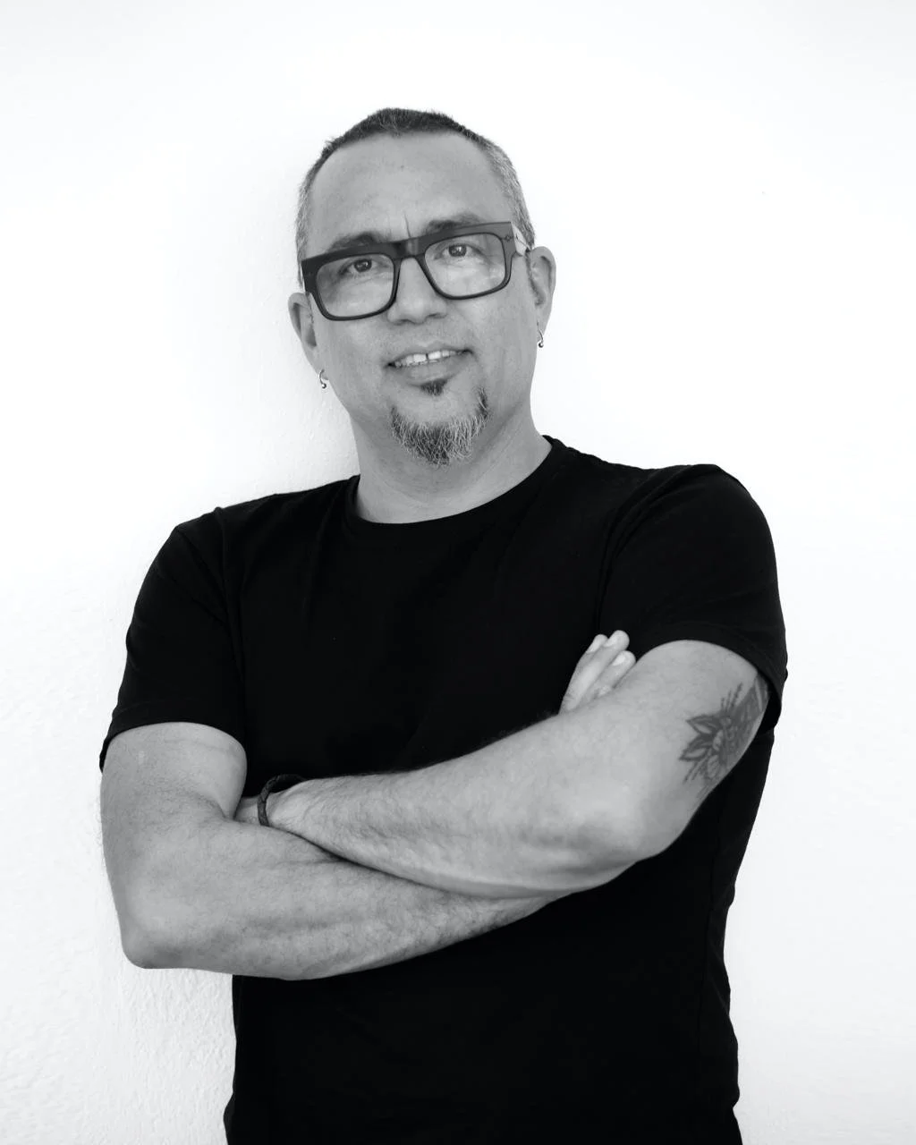 Retrato de Salva Carbó en blanco y negro de un hombre con gafas, barba y tatuajes, sonriendo y con los brazos cruzados, de fondo blanco.