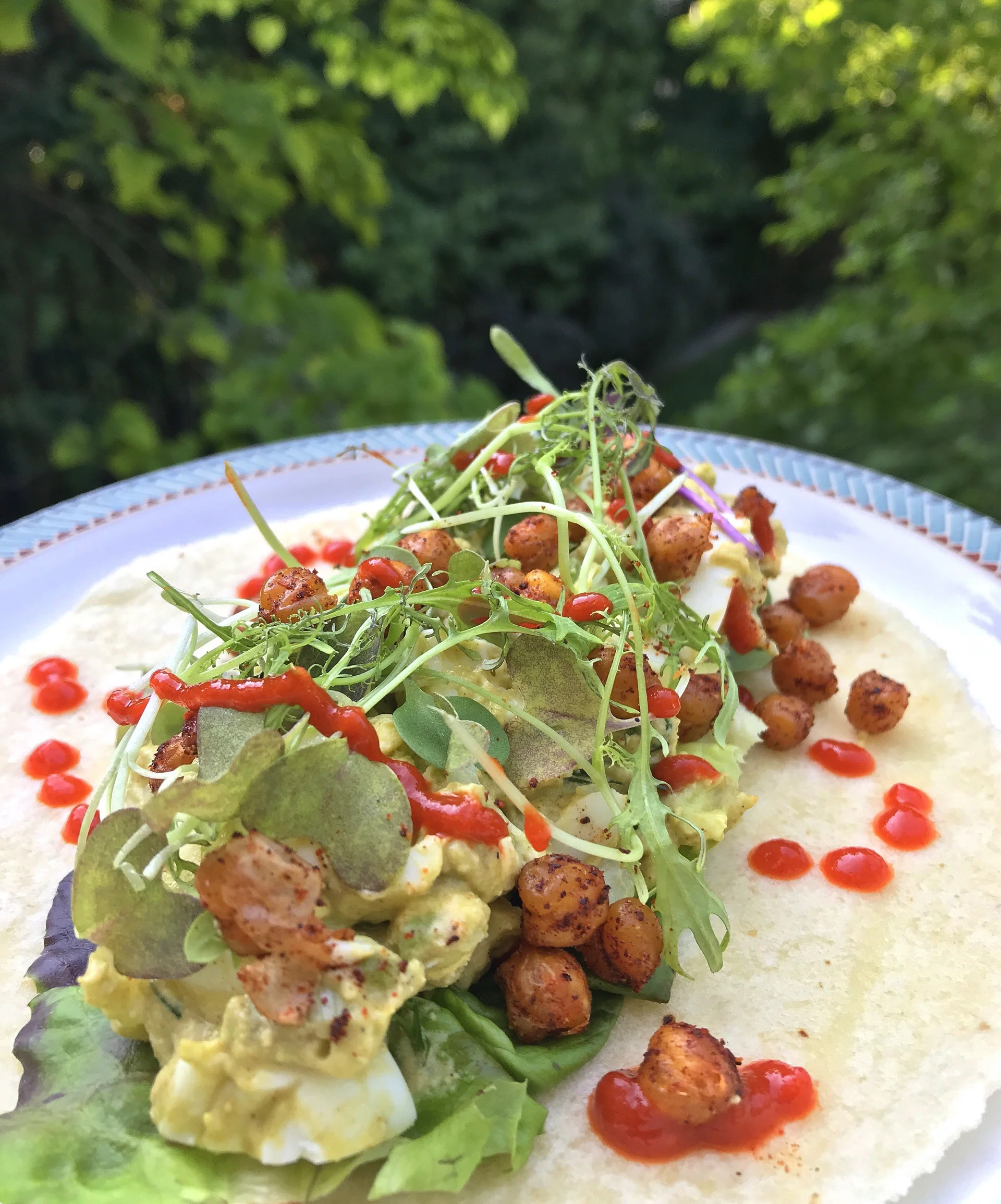 Avocado Egg Salad with fresh coriander served taco-style on a grain-free almond flour tortilla with baby lettuce, microgreens, spicy roasted chickpeas, and sriracha.