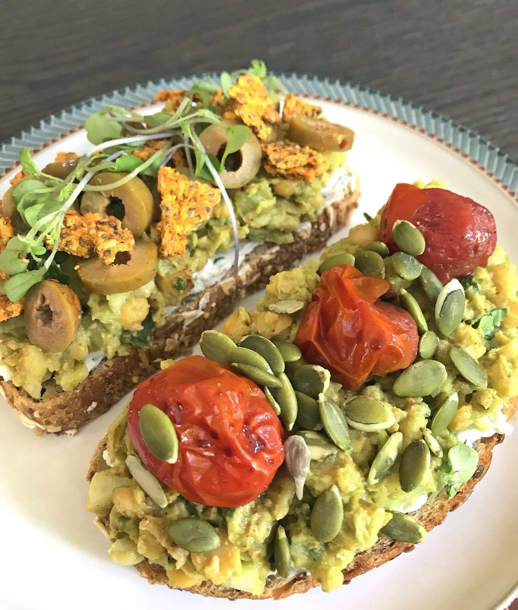 Smashed Chickpea & Avocado Salad served on toasted sourdough with vegan almond milk cream cheese, and (L) diced spicy olives, microgreens, and vegan BBQ crackers, and (R) roasted grape tomatoes and pumpkin seeds.