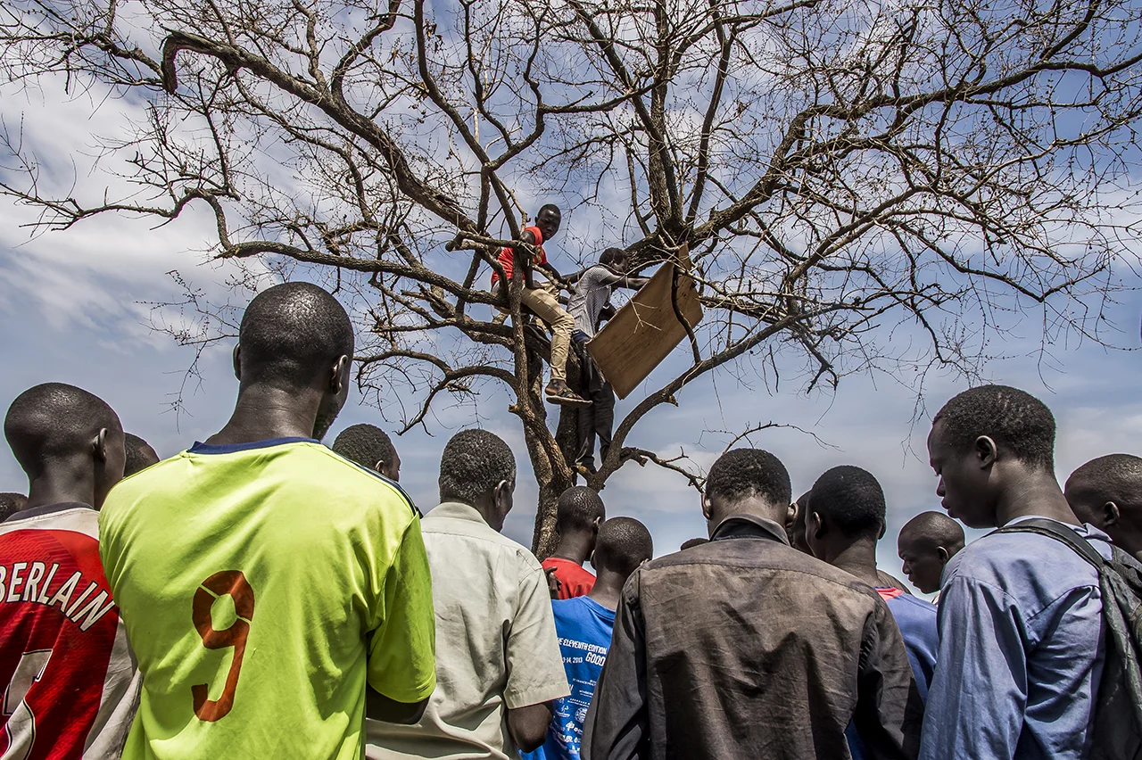 Imvepi reception Center - In the morning, a crowd of refugees gathered around a tree, where two men have climbed. These men call the names of refugee families, using a list based on the registration made by the head of families in the days before, and established by Office of Prime Minister (OPM) staff. When their name is called, the refugee can climb in a truck and load their belongings in a luggage truck. They will then be driven to an allocated plot, somewhere in Bidibidi where a new life is supposed to start.