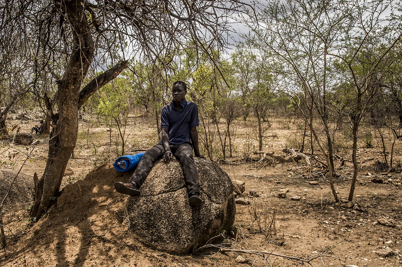 Bidibidi settlement area. A refugee sits on a rock located on the plot of land allocated to him. He was dropped off with other families by trucks along the road, with his belongings nearby. South Sudanese refugees are on land lent by Ugandan peasants. Upon arrival, they receive a blanket, soap, a blue plastic mat and a can. The essential jerrican is used to collect water from large tanks, which are supplied twice daily by tanker trucks. The quality of water, made safe by the addition of chlorine, is monitored twice a day by NGO-trained refugees.