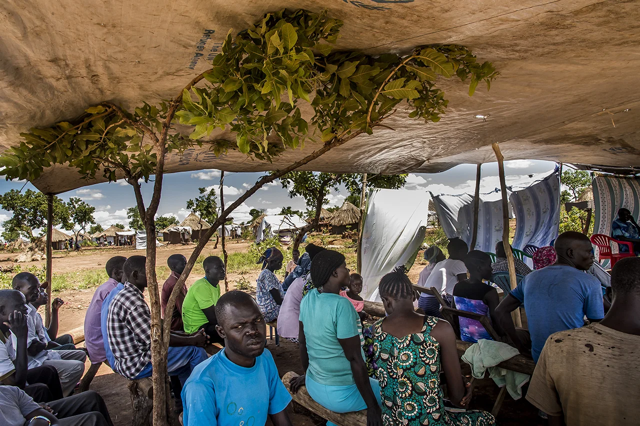 Last praying ceremony for Lucia TOKOSANG, born on 10 July 1951. She died of heart failure after having gotten malaria. The ceremony is held in Lucia's family concession and is presided by the Reverend Jephania Fongale Ezibon, the Anglican pastor of Bidibidi (priest since 1978), a south Sudan refugee. The relatives gather under the shade of a canvas to listen to tribute to the deceased.