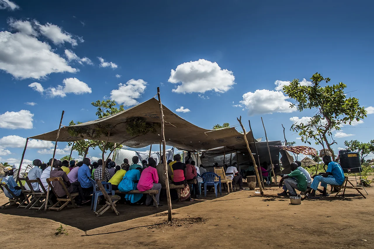 Last praying ceremony for Lucia TOKOSANG, born on 10 July 1951. She died of heart failure after having gotten malaria. The ceremony is held in Luci's family concession and is presided by the Reverend Jephania Fongale Ezibon, the Anglican pastor of Bidibidi (priest since 1978), a south Sudan refugee. The relatives gather under the shade of a canvas to listen to tribute to the deceased.