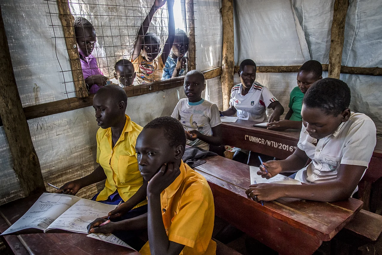 Koro Highland Primary school one out of the 30 primary school existing in Bidibidi settlements. This is a day of examination. Young pupils fill their examination sheets, while some younger one watch upon from outside, leaning on the window, a wire netting. The classroom is a long white UNHCR tent, with table provided by the UN refugee agency. The number of pupils is 2883. There are officially about 65 Ugandan pupils, but teachers say that the real figure is more than 200. Some Ugandan fear to confirm there nationality, being afraid to be expel from the school, as they think that it is reserved for South Sudanese only.