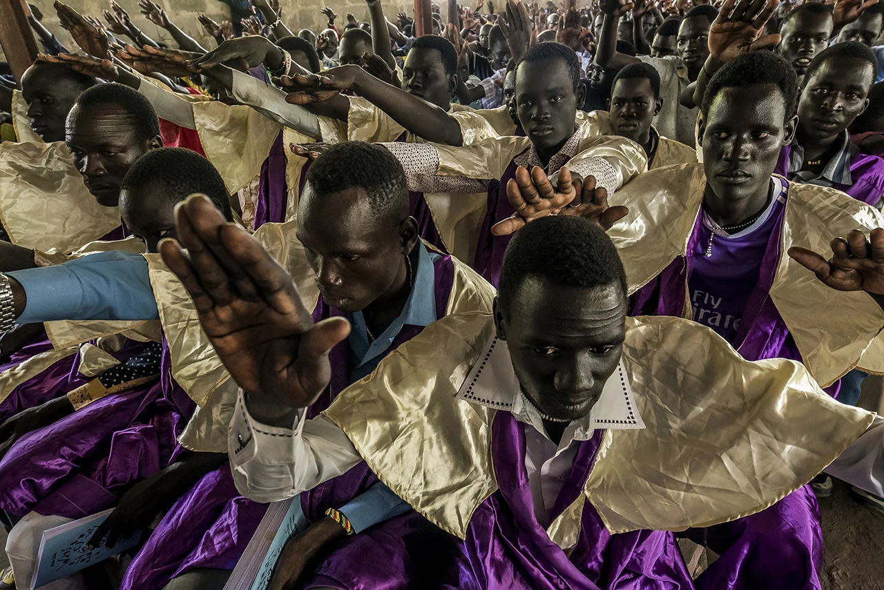 A group of young men respond to a deacon's announcement during Sunday morning religious service in Old Fangak's St. Daniel Camboni Catholic Church, run by the Comboni Missionaries, waving their hands over their heads, cheerfully.