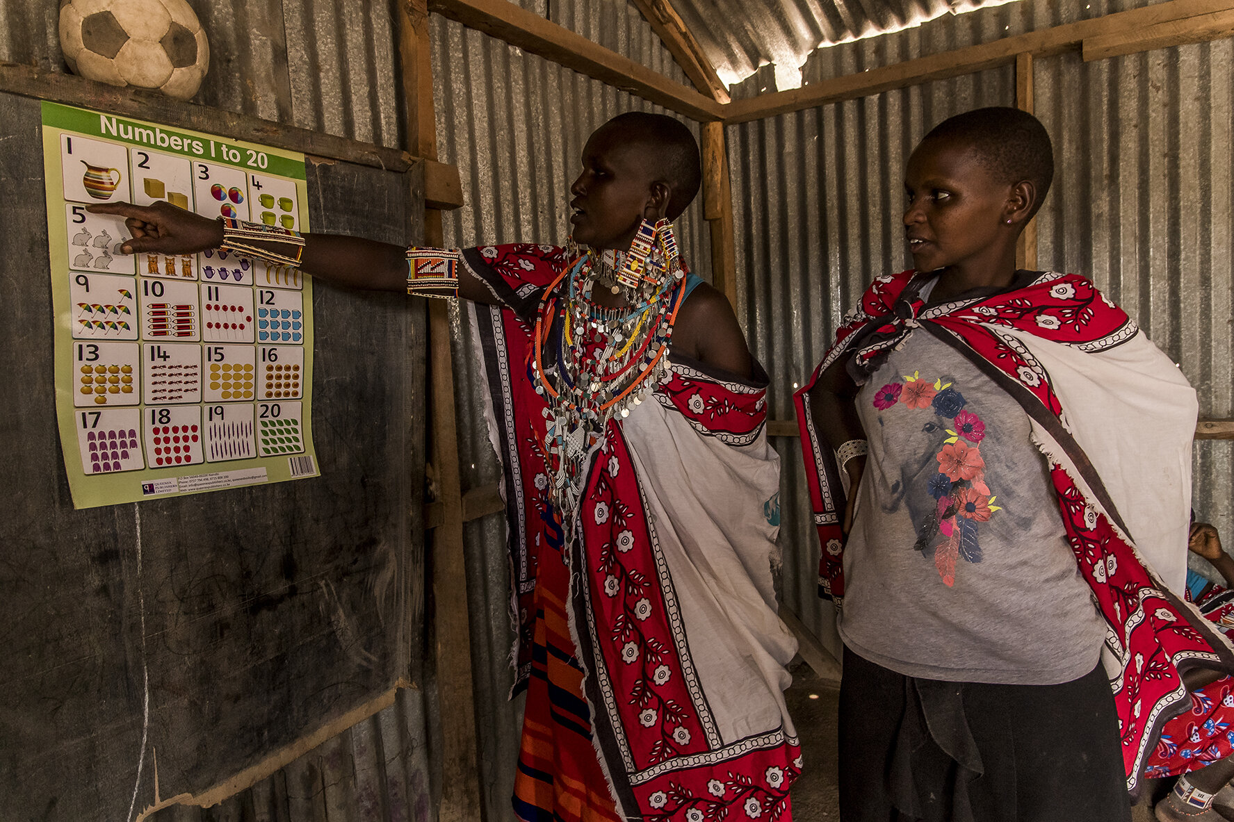 A young Masai woman teaches a member of her tribe to recognize numbers, in the school building built by women and financed in particular by the sale of handcrafted jewelry that they make. Une jeune Masaï apprend à une membre de sa tribu à reconnaît