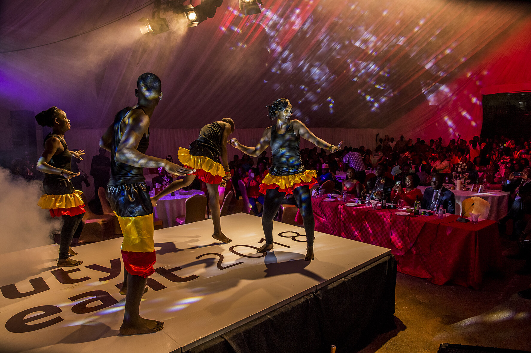 The beauty pageant begins with a choreography bringing together all the candidates, boys and girls, in front of a packed marquee (gazebo). They wear all the colours of the Ugandan flag: black, red and yellow. The members of the jury are seated in th