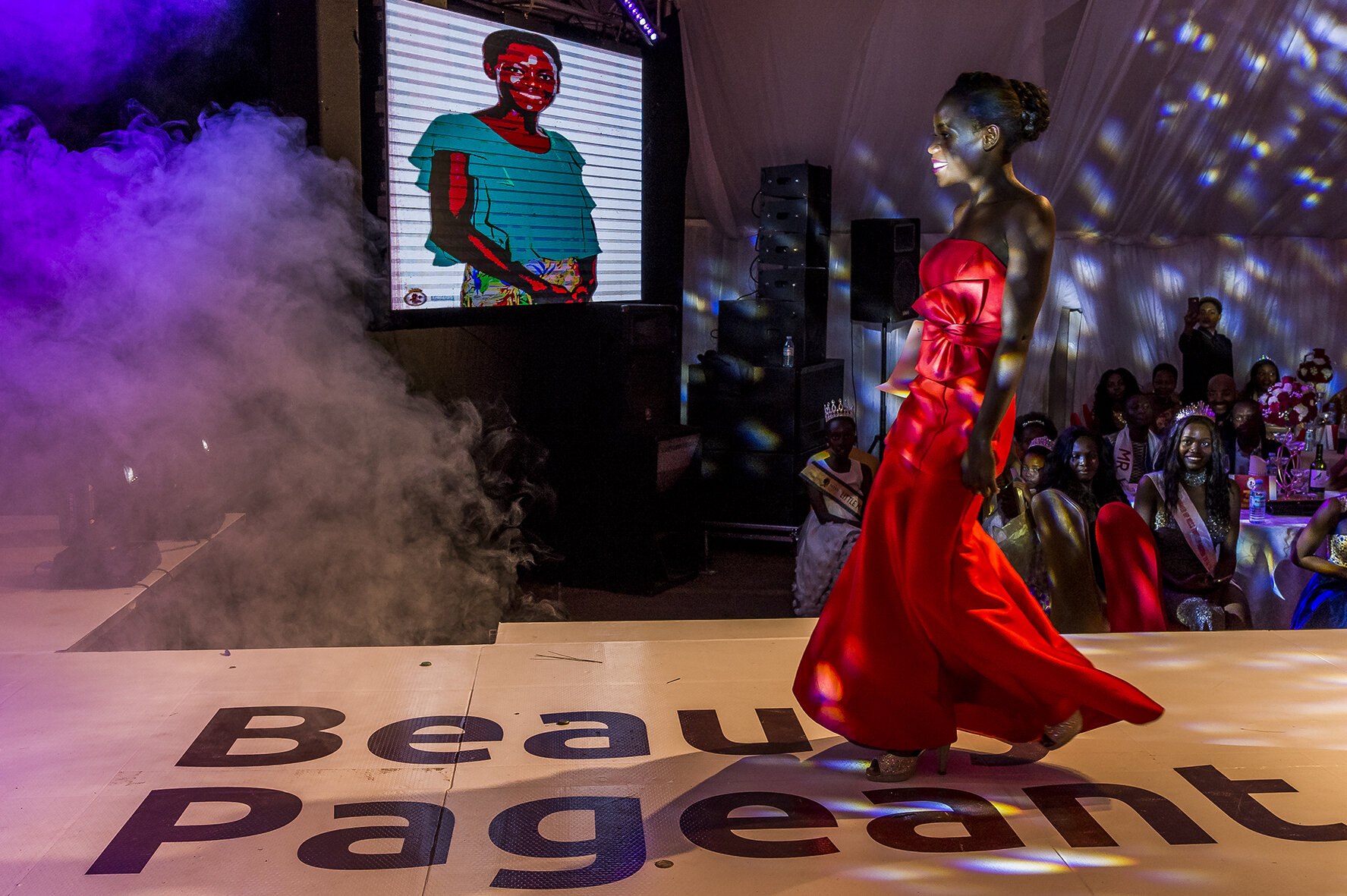 A candidate wearing an evening dress makes her way on the podium before the five Ugandan jurors. This is one of the beauty contest tests. On the giant video screen, her portrait is displayed while in the room a mistress of ceremonies presents her.