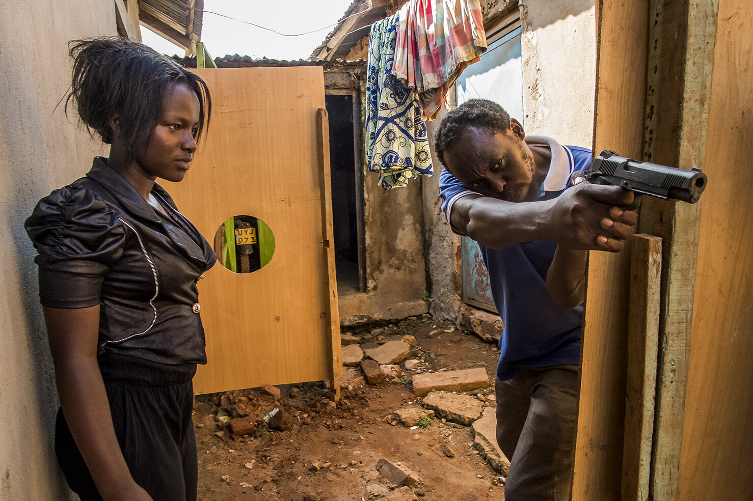  In Wakaliga slum, Issac shows Ritha, how he wants her to use her gun, for a scene shot in a jail decor, built in his courtyard. She is the first ever heroine of Wakaliwood, the main character in « Who killed Captain Alex » a TV series, inspired from