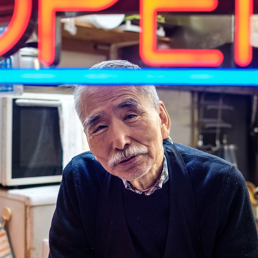 Portrait of an elderly Japanese man in front of a glowing neon OPEN sign in Japan — street documentary photography by Oliver Maximilian, KULTURHYBRID®