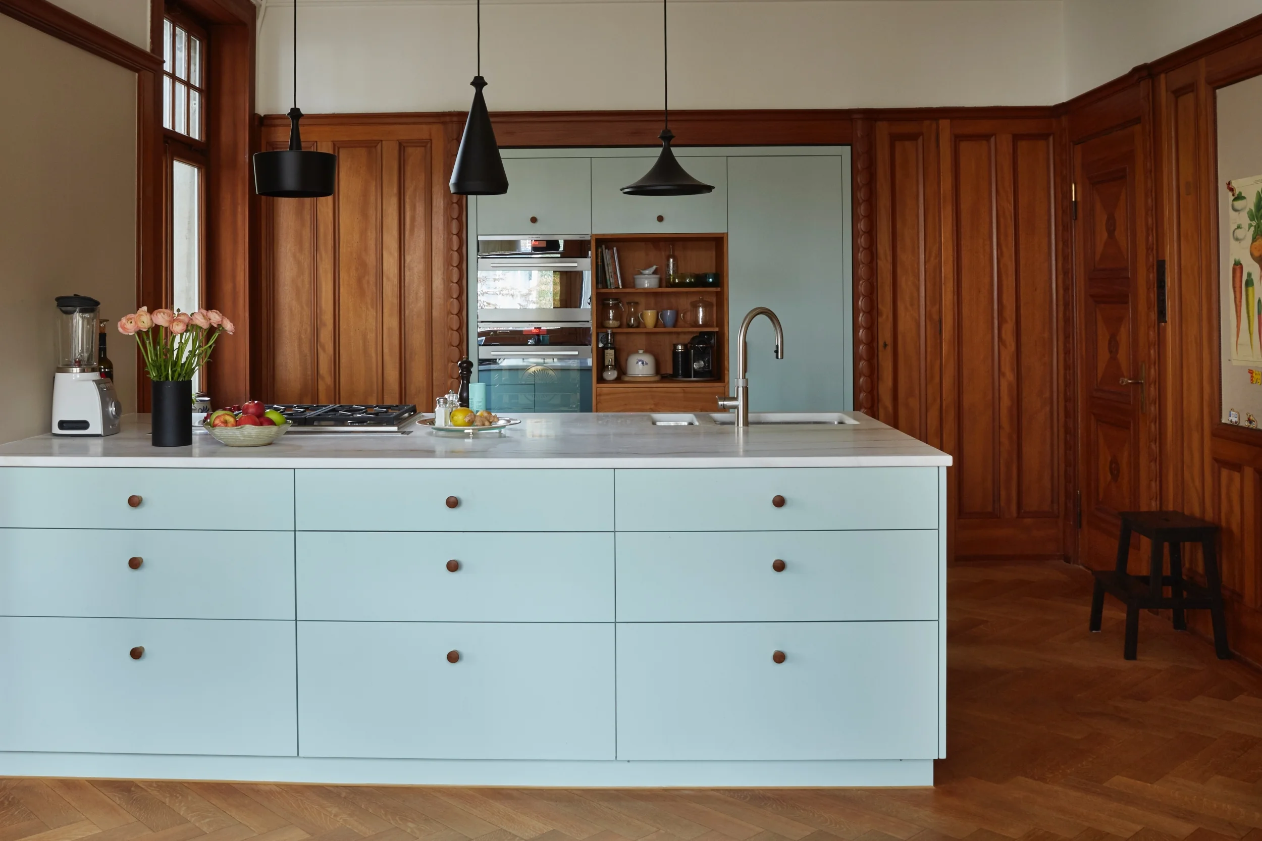 Diese Küche wurde im ehemaligen Salon der Villa eingerichtet. Neben der grossen Insel nutzten wir die Nische einer ehemaligen Verbindungstür für den Hochschrank. Das bestehende Täfer ist aus Mahagoni, aber die Griffe und das offene Regal sind aus einheimischer Kirsche gefertigt. Farblich passt es sehr gut zum exotischen Mahagoni und integriert die neue Küche perfekt in die alte Architektur. Das dezente Hellblau der Fronten macht das grosse Möbel leicht und steht in kühlem Kontrast zum warmen Täfer der Wände.
