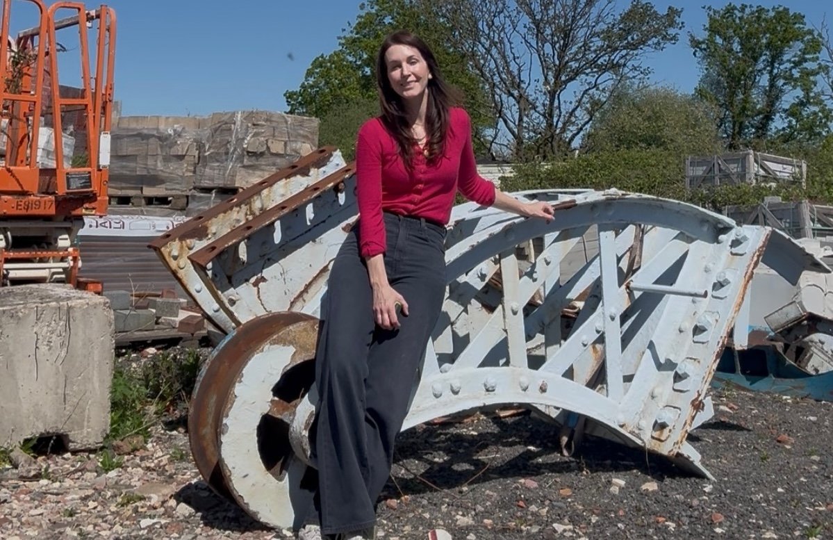 Woman leaning on large irregular shaped salvage metal in an outdoor location. Machinery and bricks around. The sky is blue and it's a sunny day.