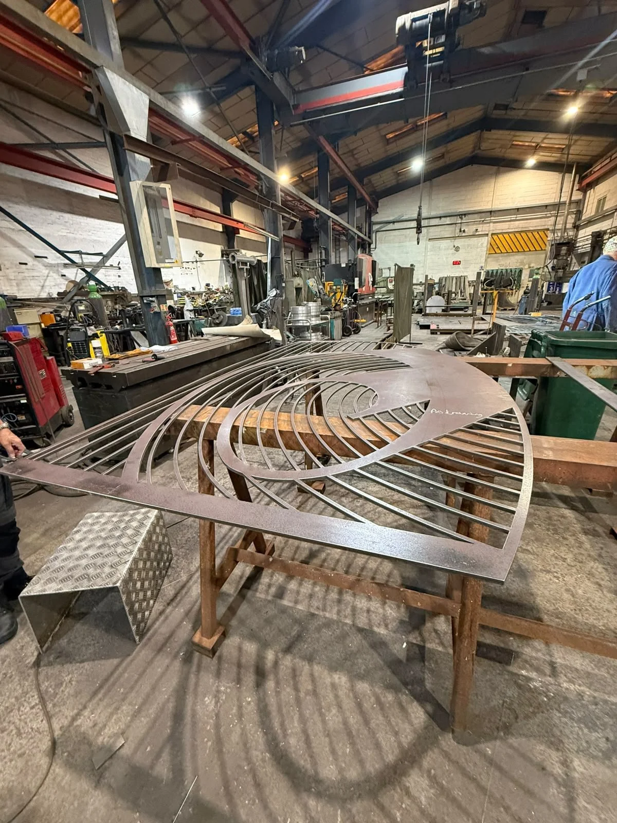 Large metal flat structure with lines and spirals laying on a workshop table, people working around it.