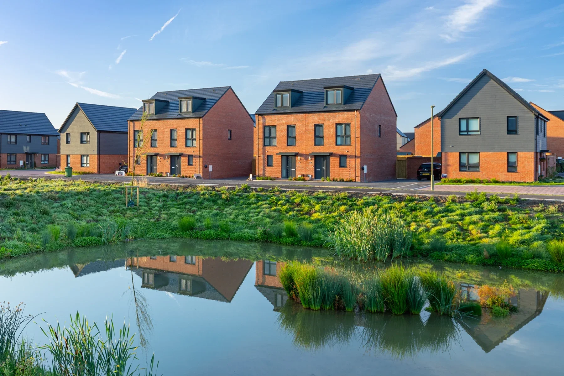 Na row of new houses, red brick and grey, behind a quiet road and pond with grass and greenery around it.