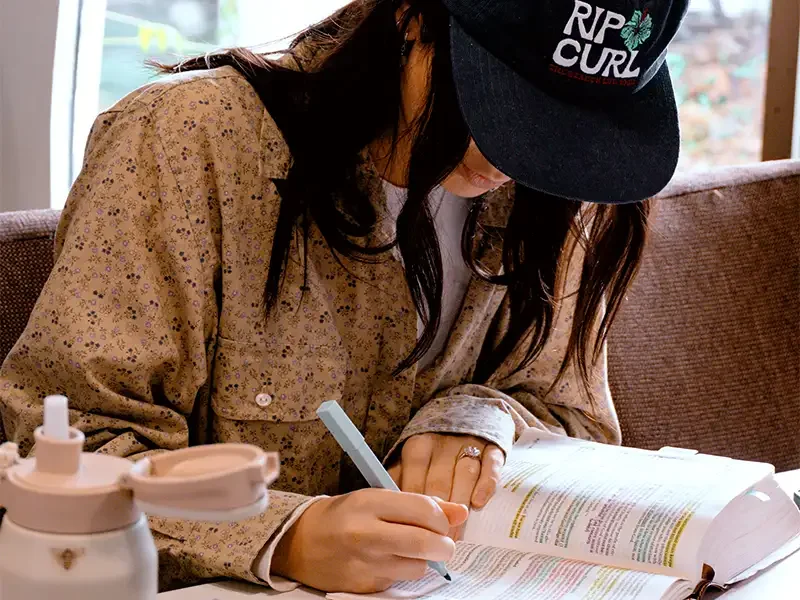 Woman with long dark hair wearing a black cap and a beige floral shirt, sitting on a brown couch, studying or reading a large open book while holding a pen in her right hand.