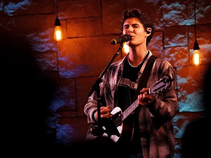 A young man with dark hair singing into a microphone while playing an acoustic guitar, on a dimly lit stage with warm orange hanging lights and a textured brick wall background.