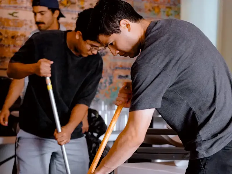 People cleaning a floor with brooms in an indoor setting.