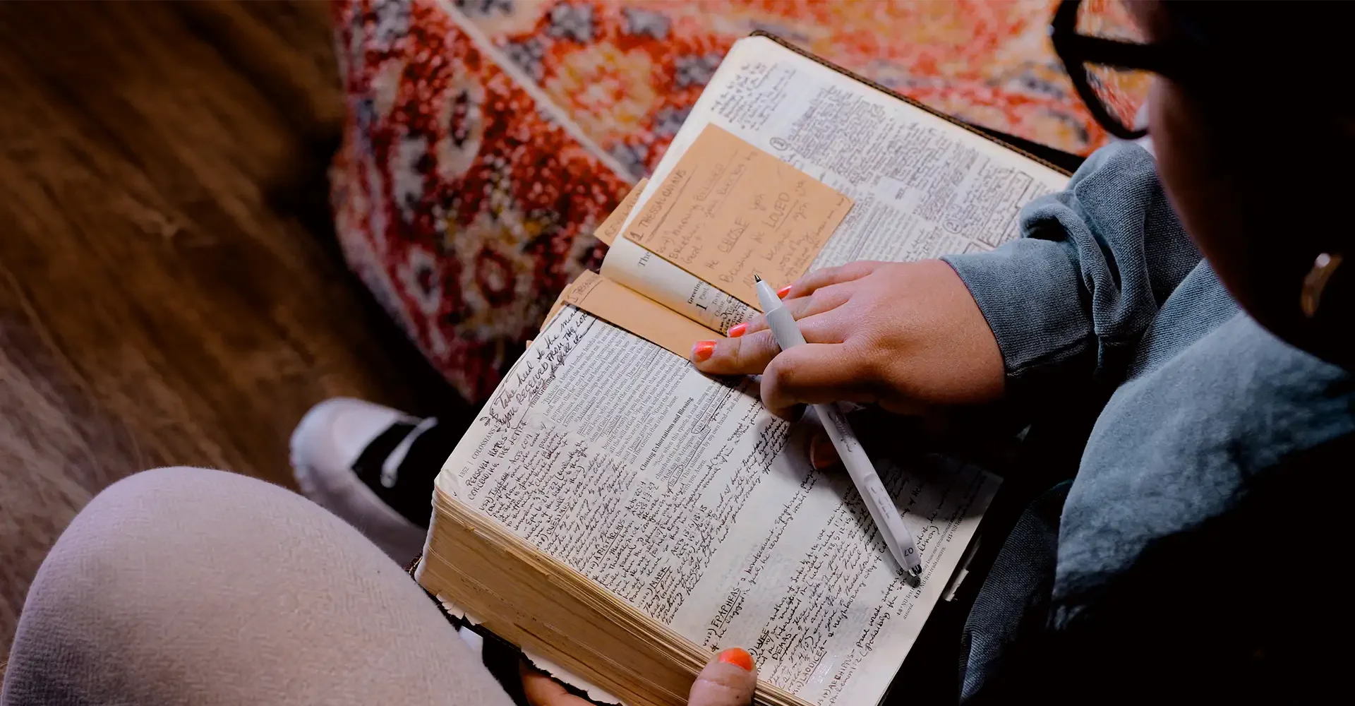 Person writing notes in a handwritten journal while sitting on a carpeted floor, with a pair of legs visible in the foreground.