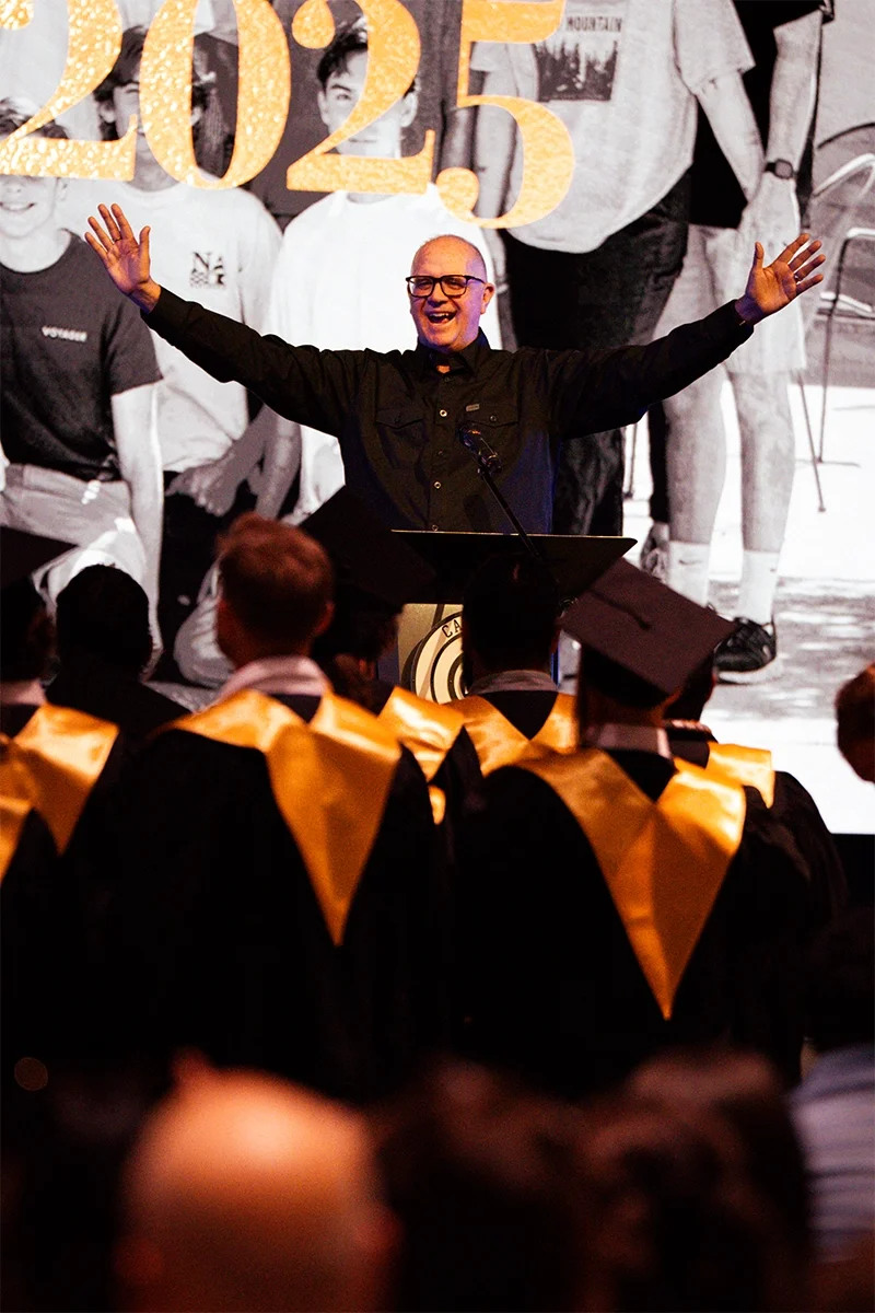 A man standing at a podium with his arms raised, speaking or giving a speech at a graduation ceremony. The audience wear caps and gowns with yellow stoles. A large banner with the year '2023' hangs behind him, and a black-and-white photo collage is displayed on the screen behind the stage.